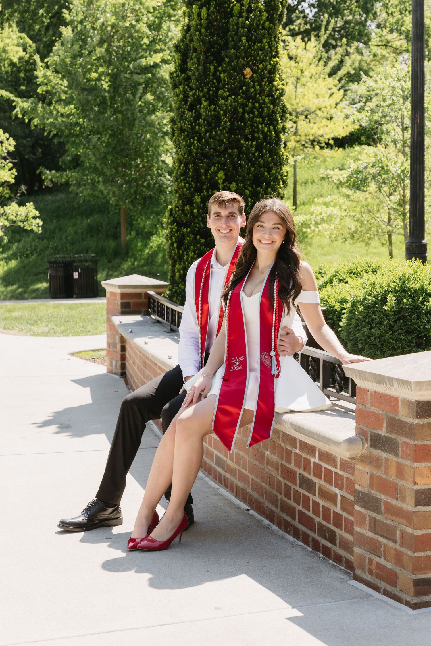 Two college graduates in red sashes pose together outdoors on a brick wall surrounded by lush green trees.