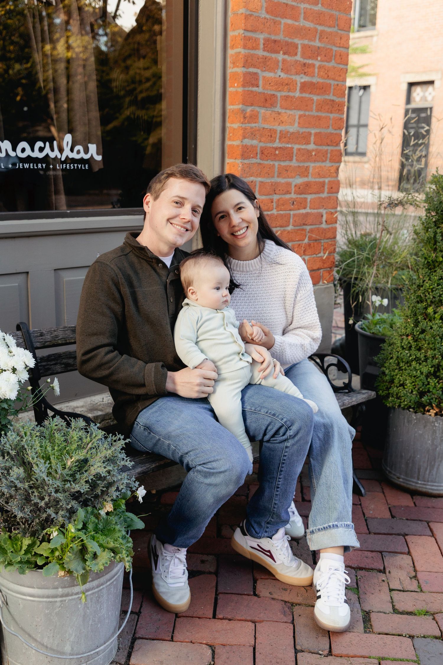 A happy couple sits on a bench outside a brick building, smiling while holding their baby dressed in a white outfit.