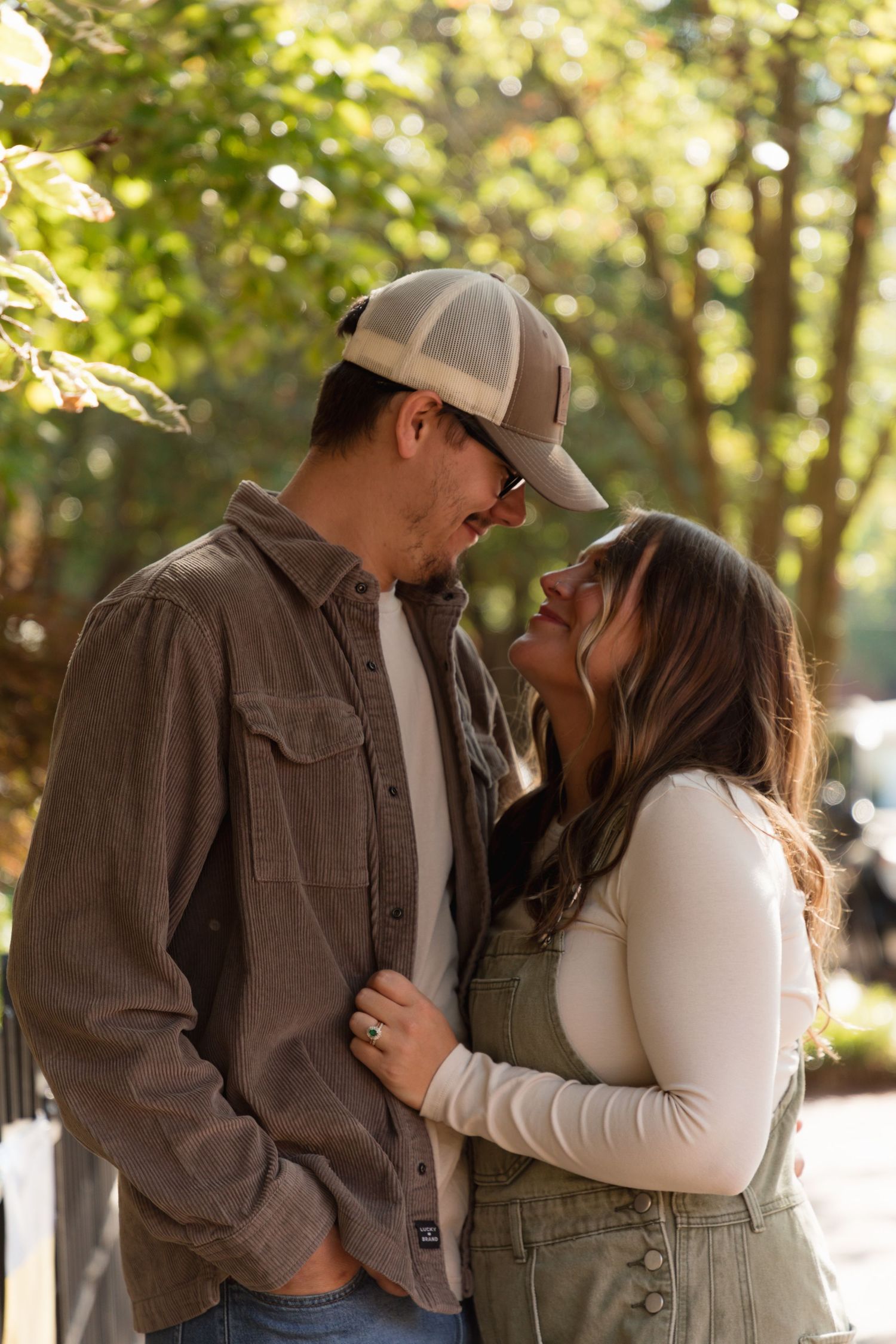 A couple in casual fall attire shares a romantic moment outdoors surrounded by green trees.