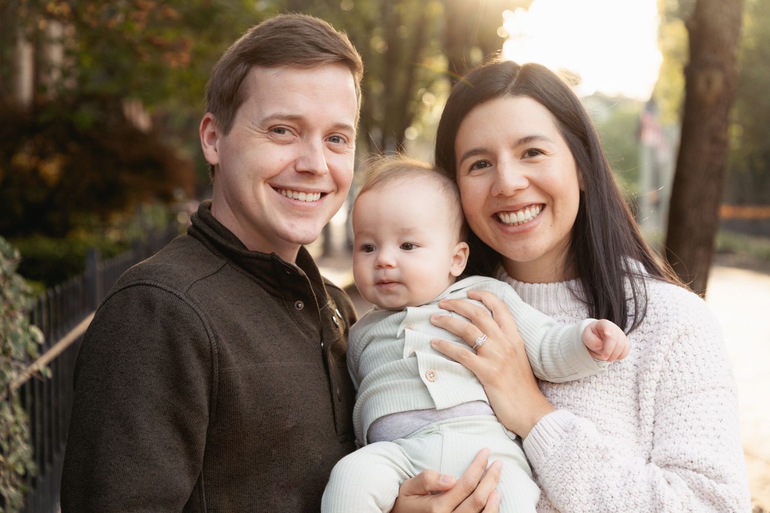 Smiling young family with parents holding their baby outdoors in a sunny park setting.