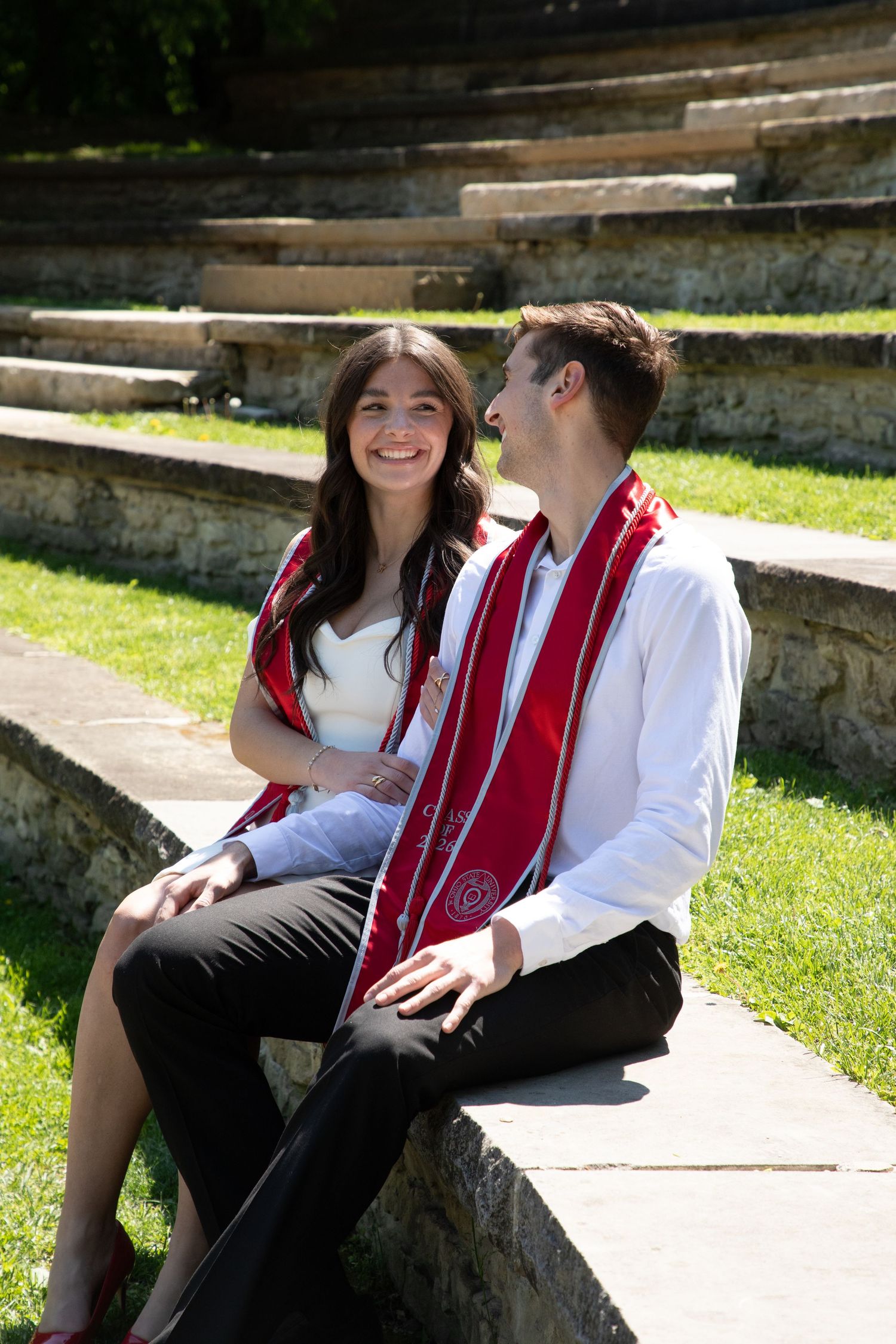 Two college graduates in red stoles sit together on stone steps outdoors, smiling and sharing a joyful moment.