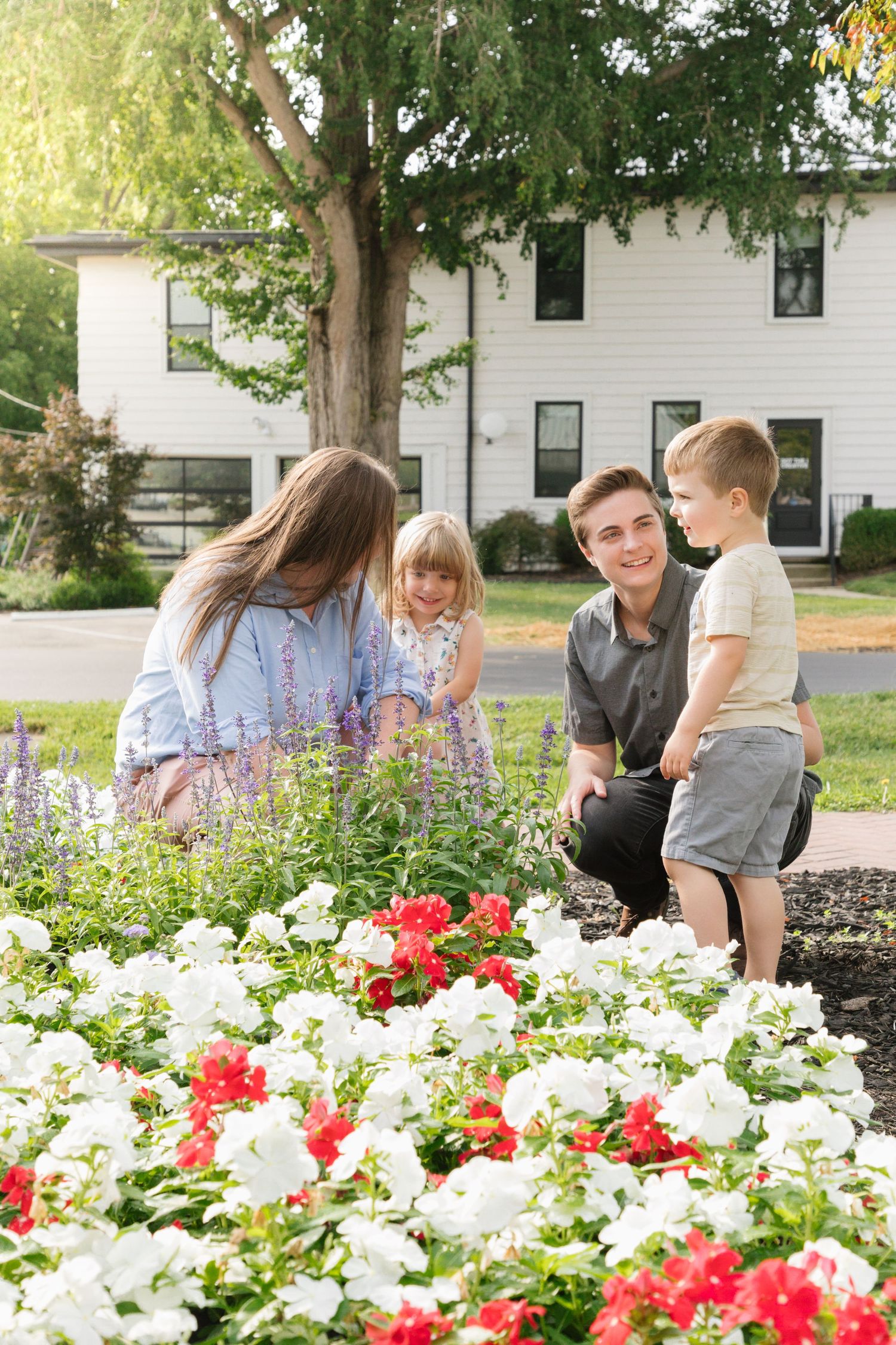 Family of four admiring colorful red and white flowers in a garden outside a white house on a sunny day.