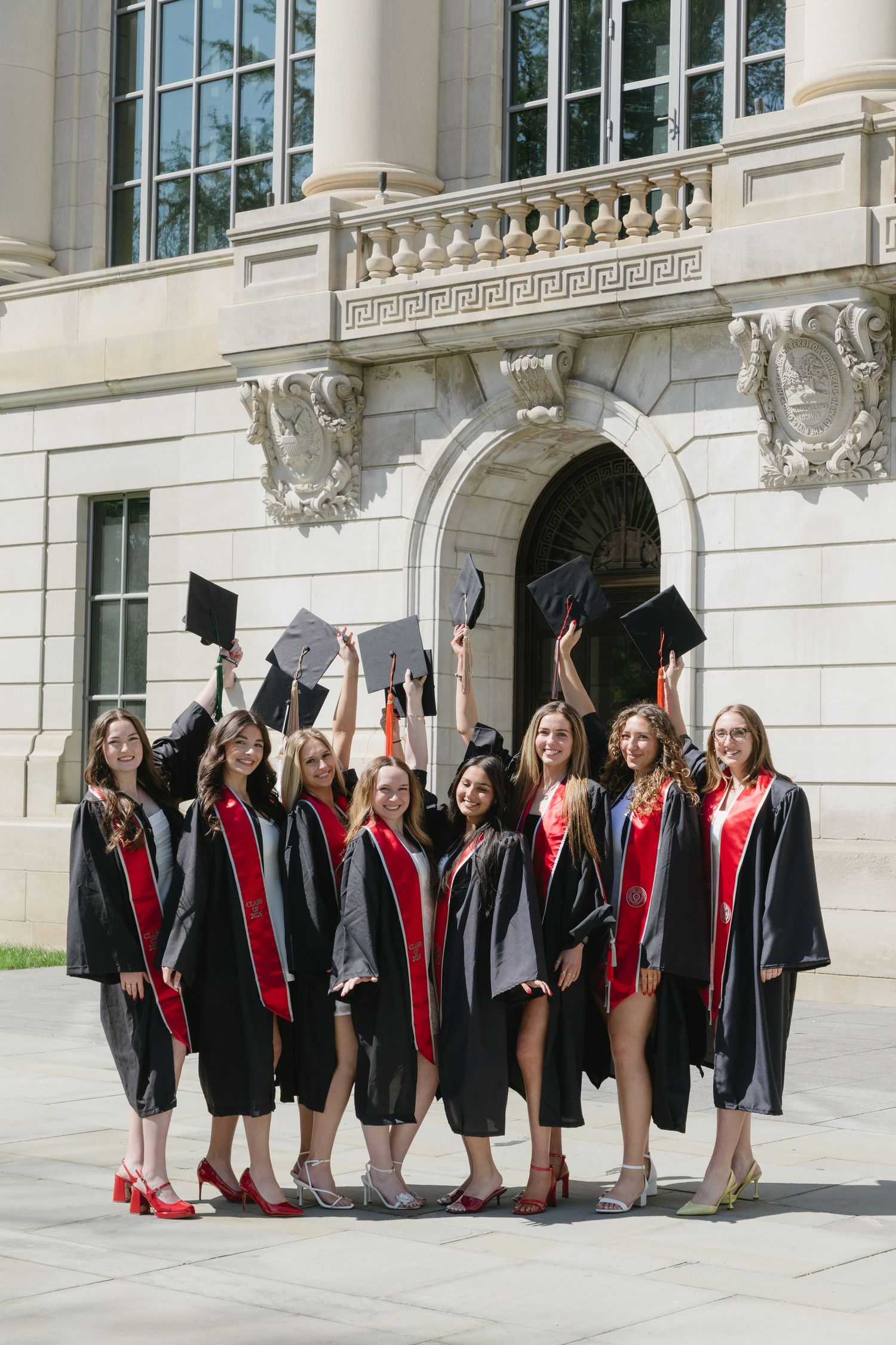 Group of female graduates in black and red gowns celebrating outdoors, tossing caps in front of a classic stone building.