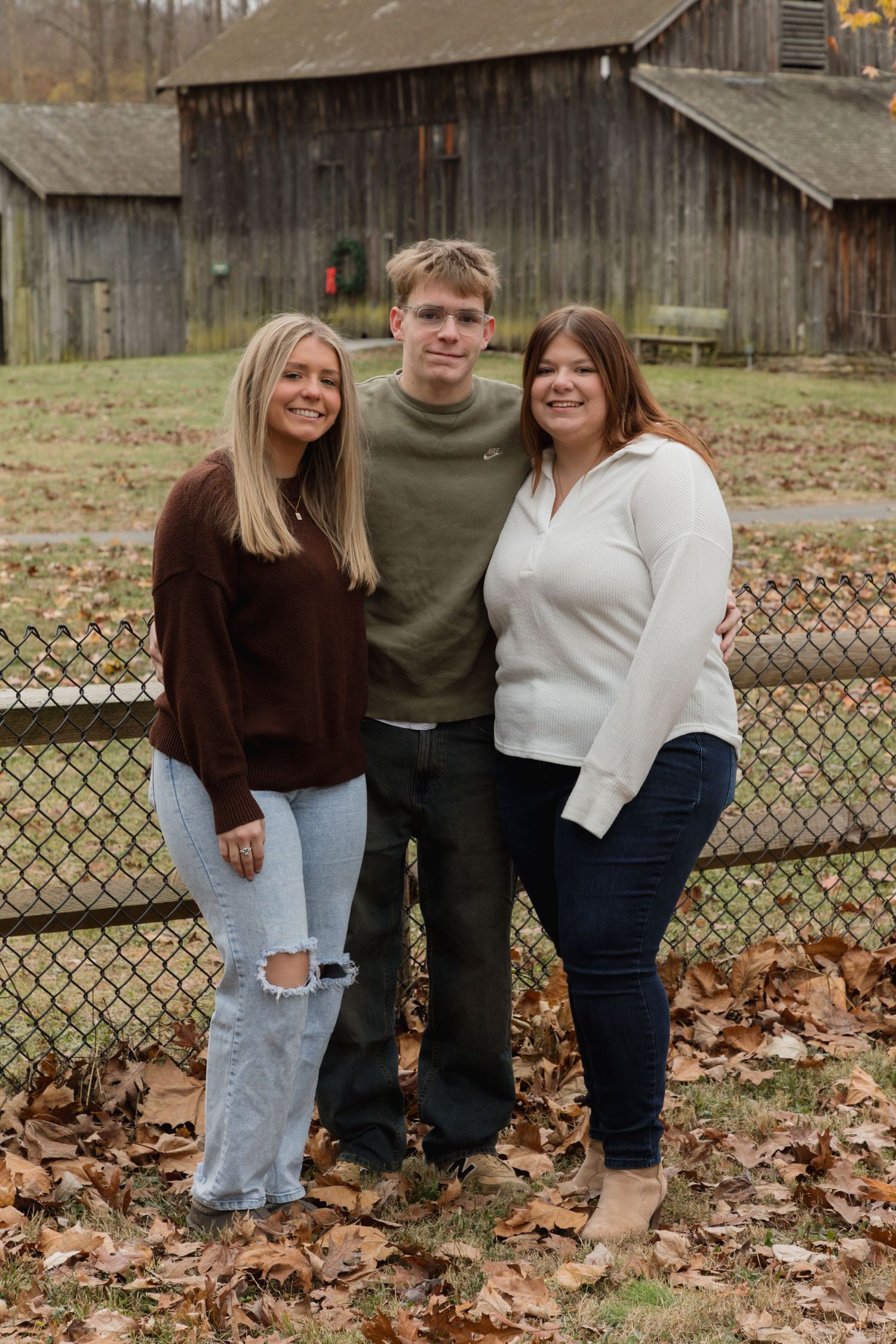 Three people smiling outdoors in fall foliage near a rustic wooden barn, wearing casual sweaters and jeans.
