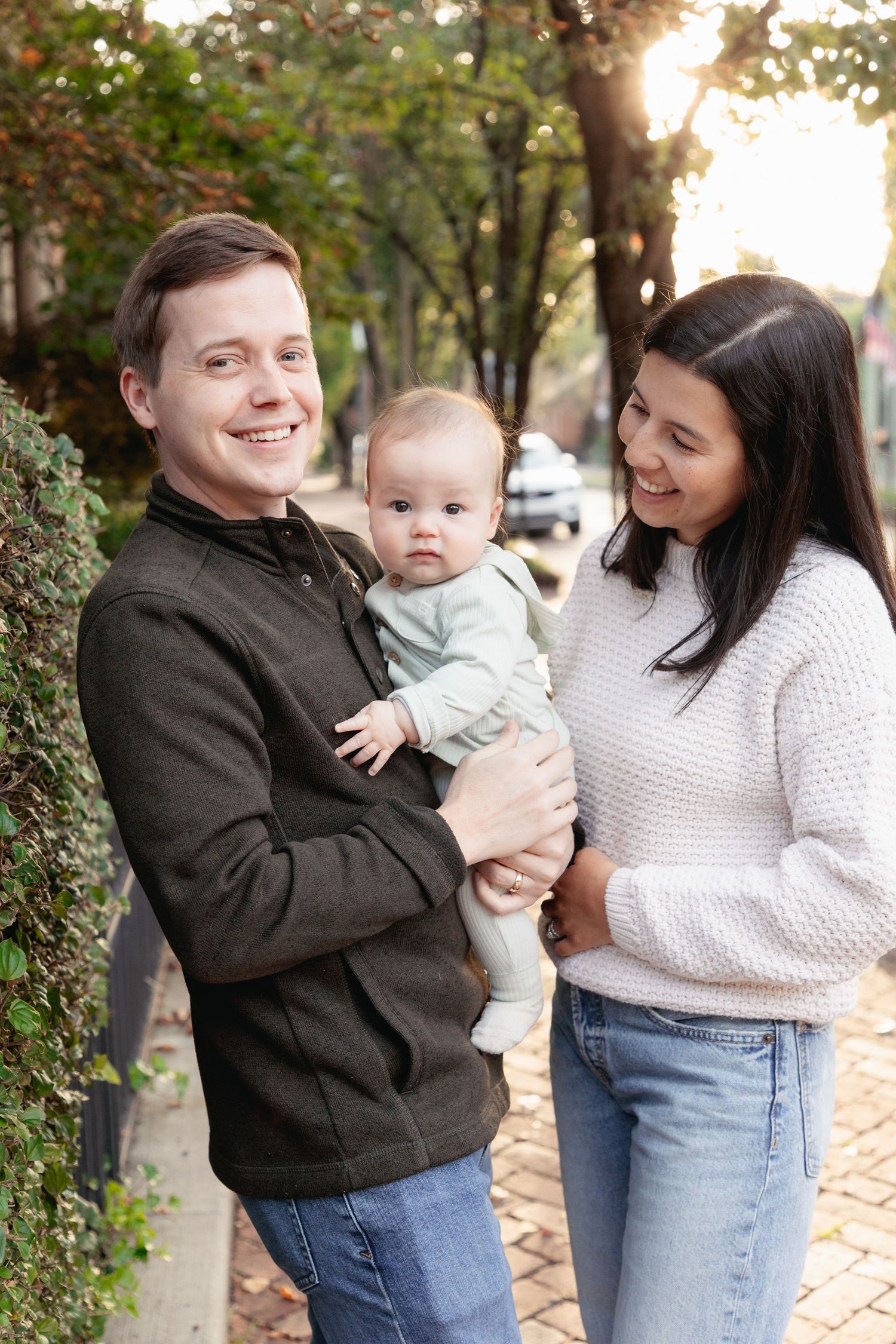 A smiling couple stands outdoors on a brick sidewalk, lovingly holding their adorable baby in a light blue outfit.