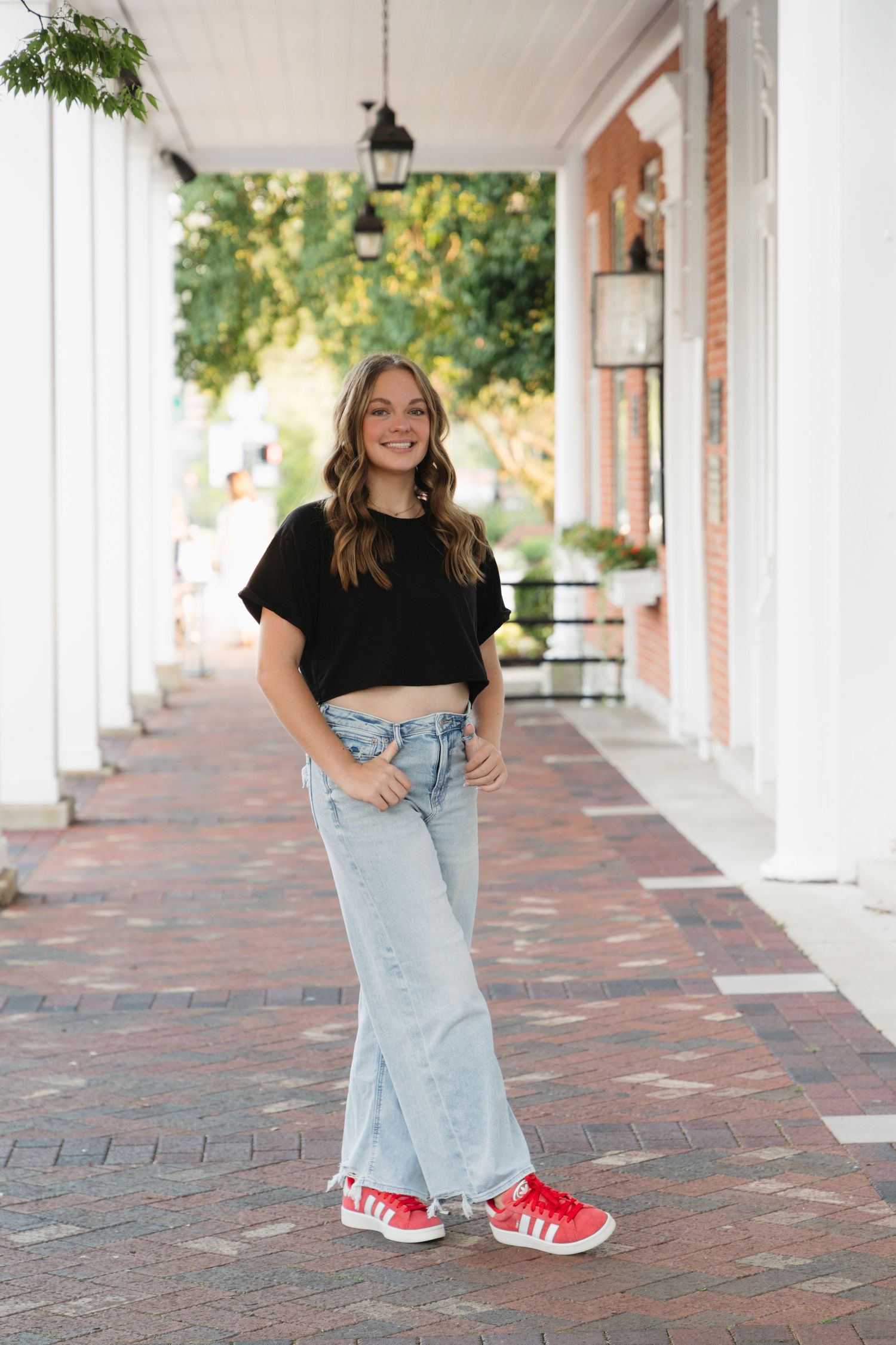 Young woman in black crop top, wide-leg jeans, and red Adidas sneakers posing on a brick walkway.