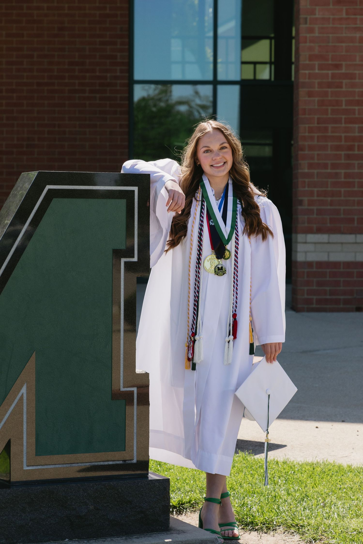 Smiling graduate in white cap and gown with honor cords and medals poses beside a large letter M outside a brick building.