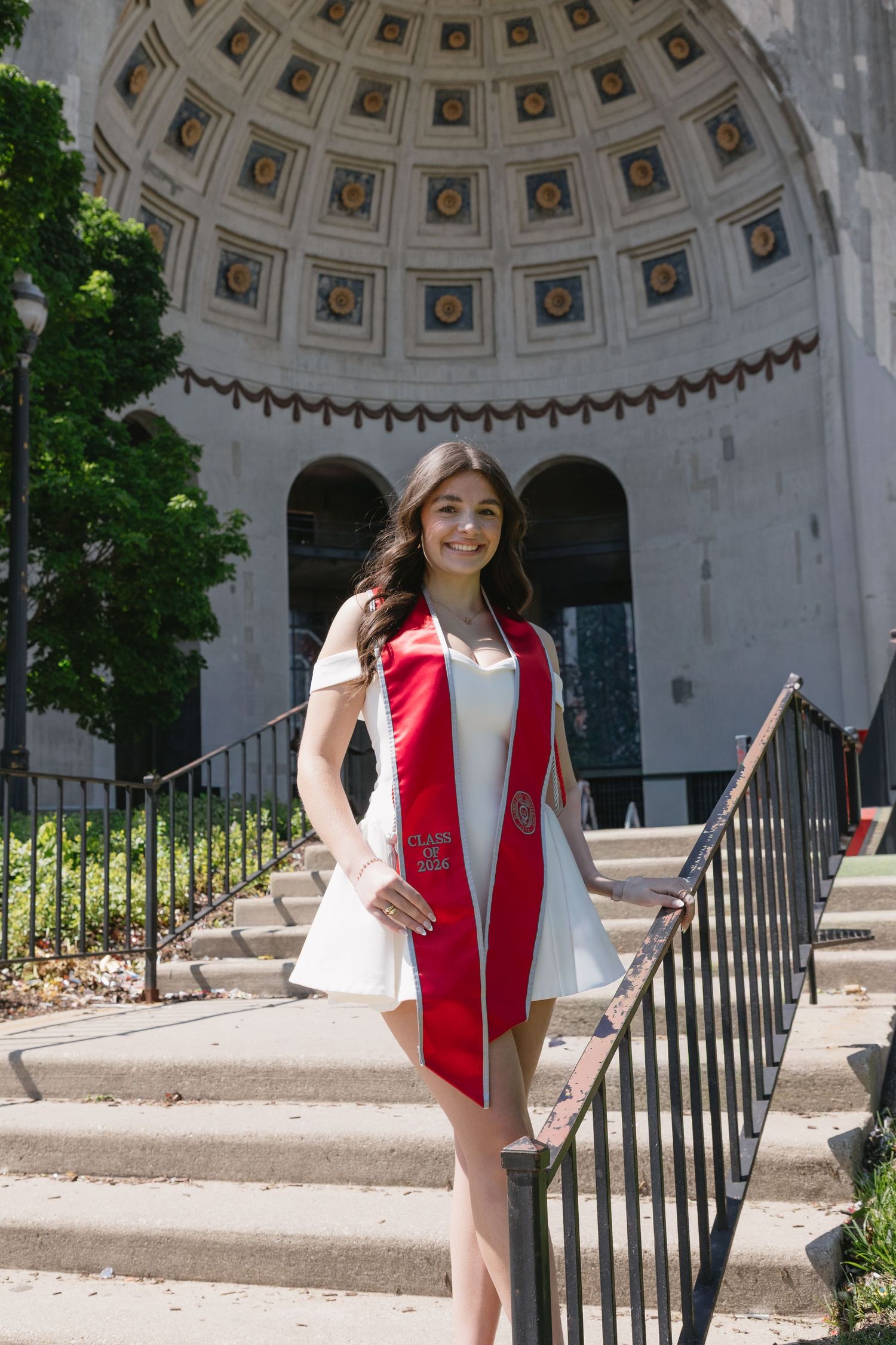 Young woman in white dress with red graduation stole poses on steps before a grand domed building on graduation day.