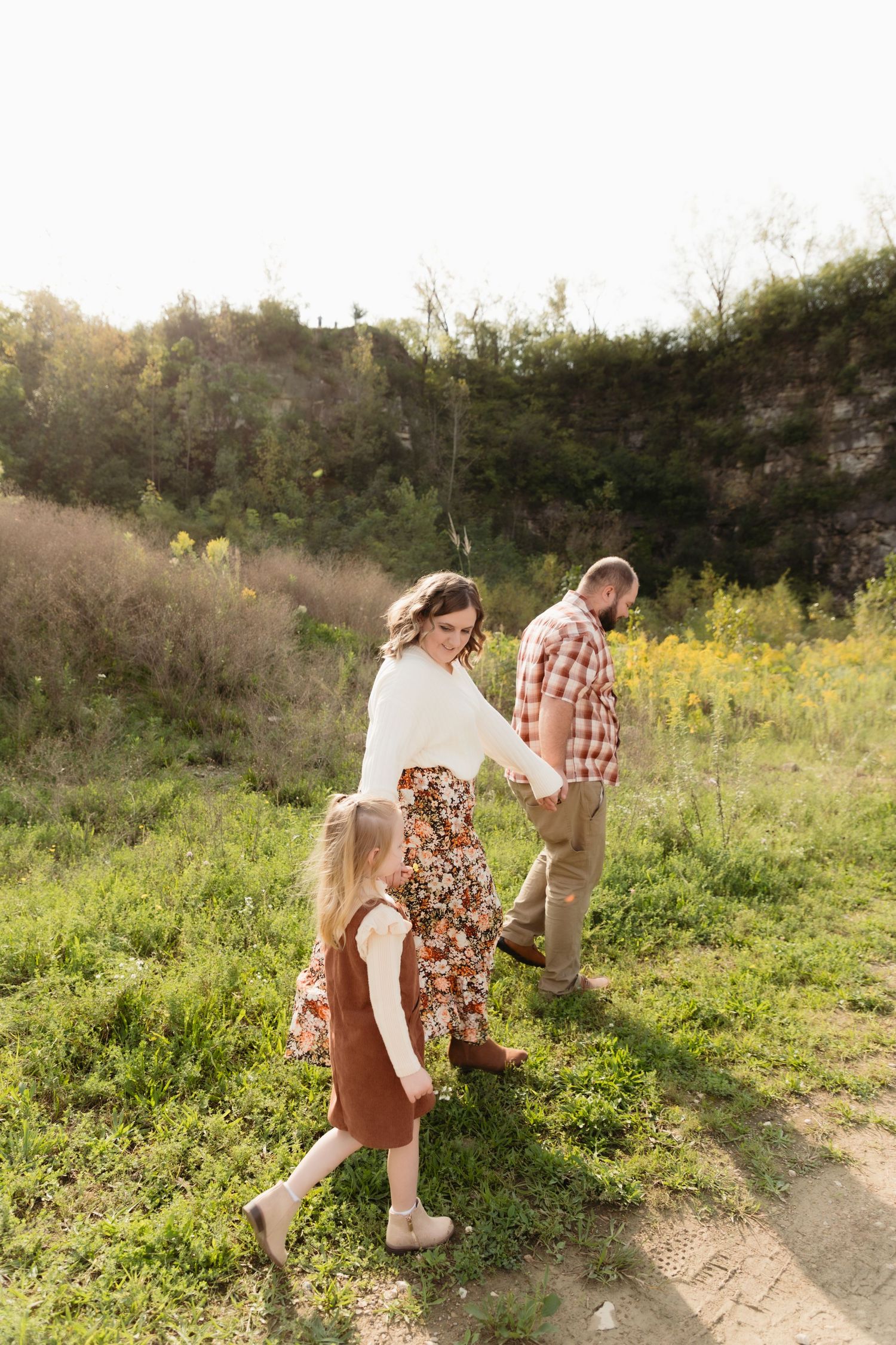 Family of three walking through a sunny meadow, mother in floral skirt glancing back, daughter in rust dress, father in plaid shirt.