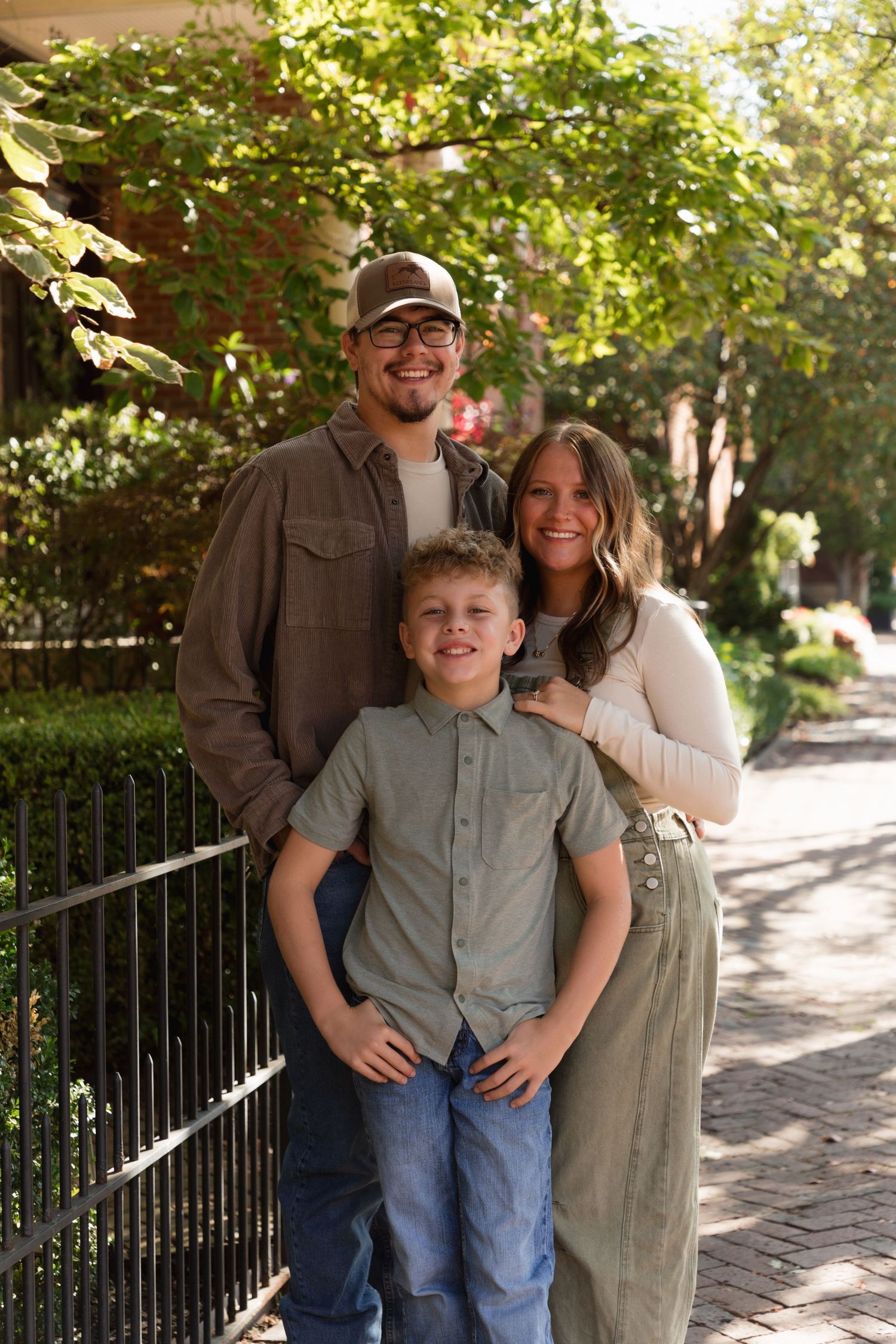 A smiling family of three poses outdoors on a tree-lined sidewalk, with a boy in front of his parents.