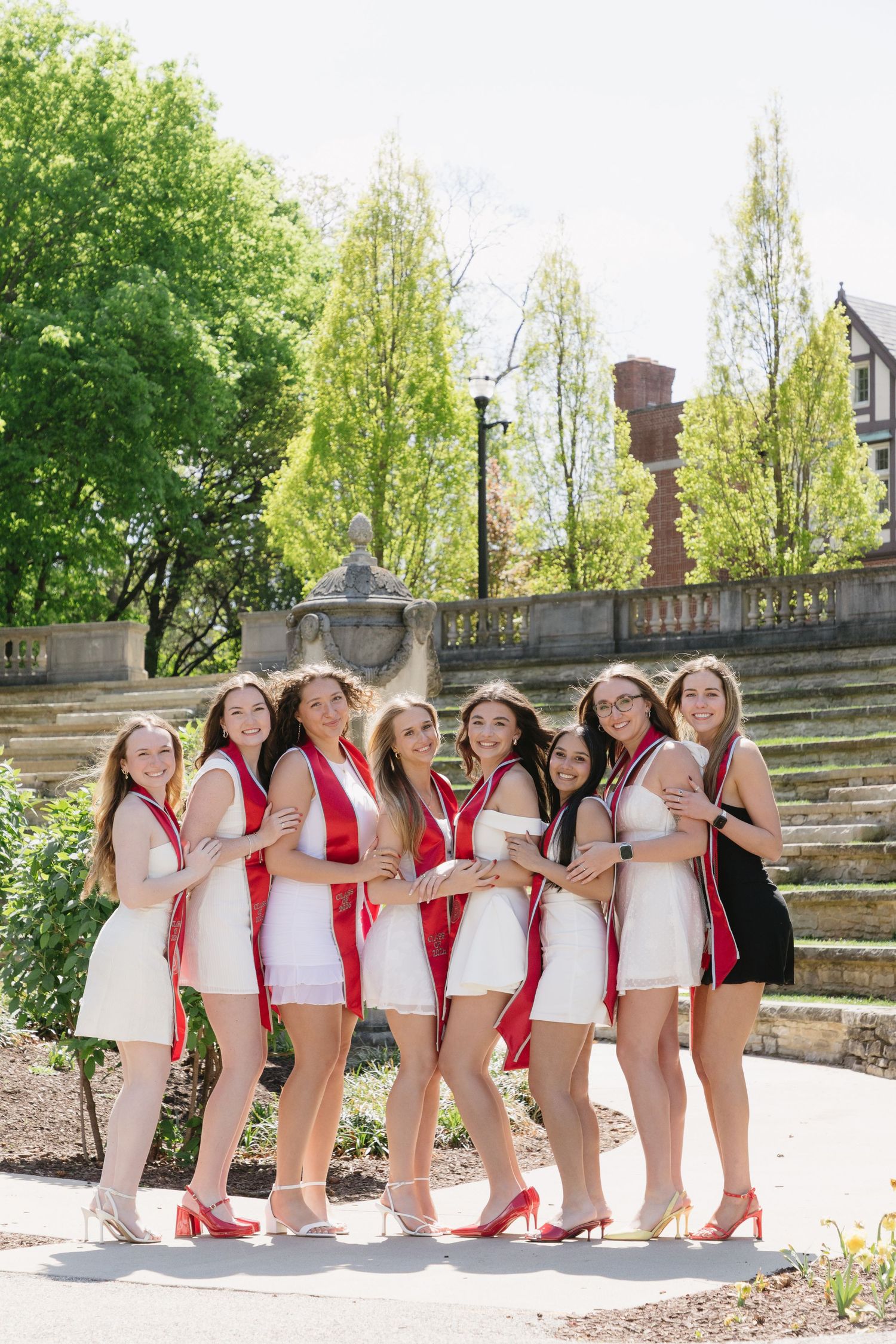 Eight young women in white dresses and red graduation sashes pose together outdoors in a park setting.