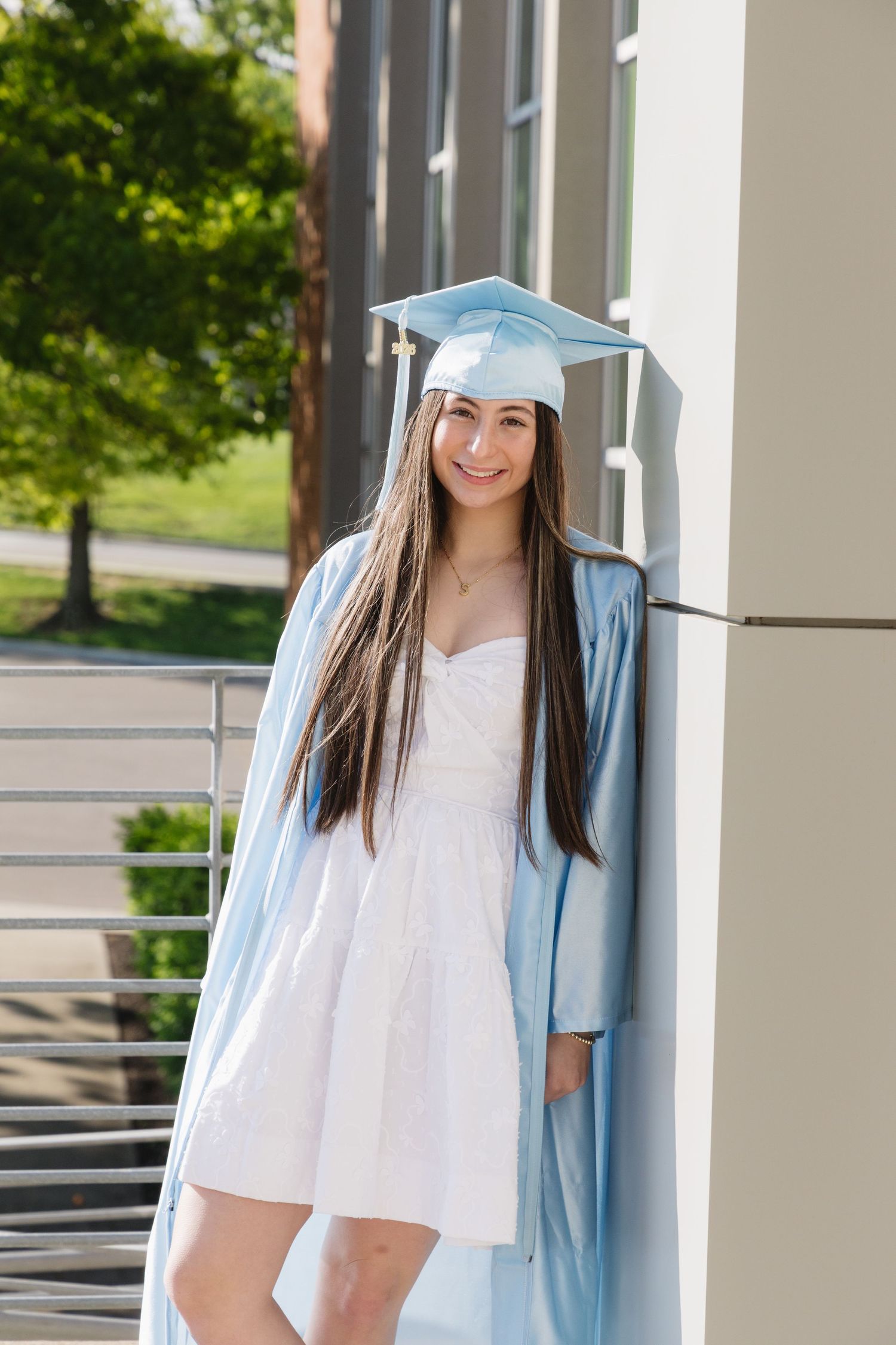 Young woman smiling in light blue graduation cap and gown over white dress, posing outdoors near building.