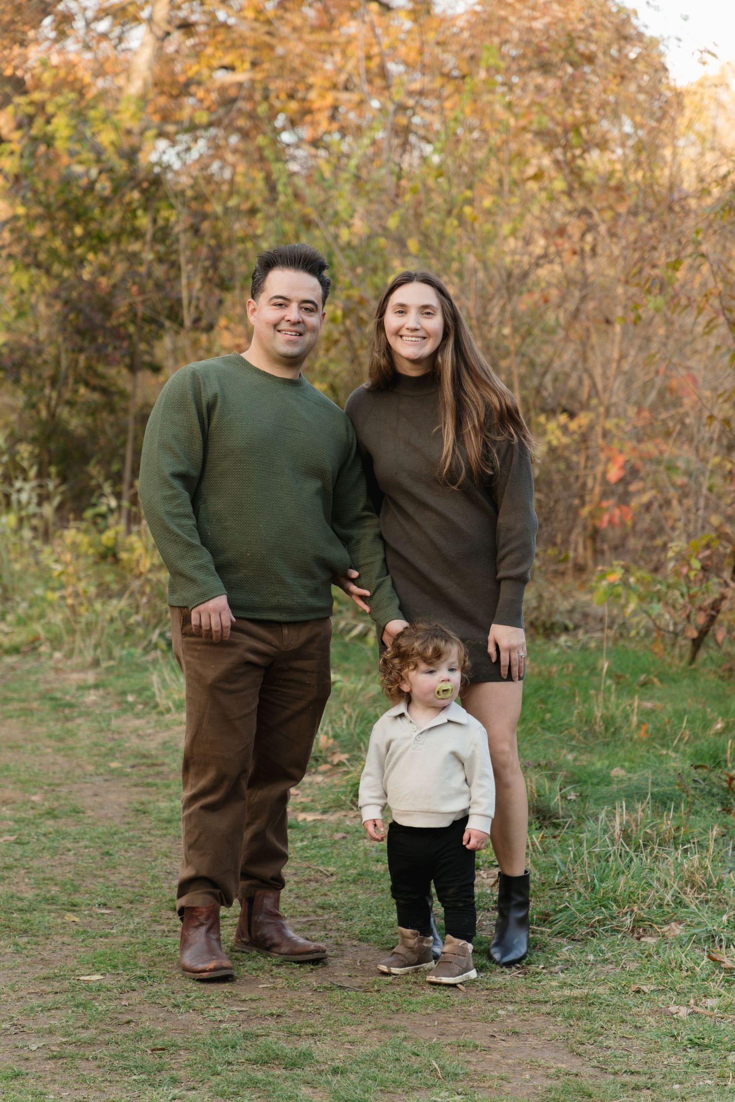 Family of three posing outdoors in autumn, parents in earth tones with toddler, surrounded by colorful fall foliage.