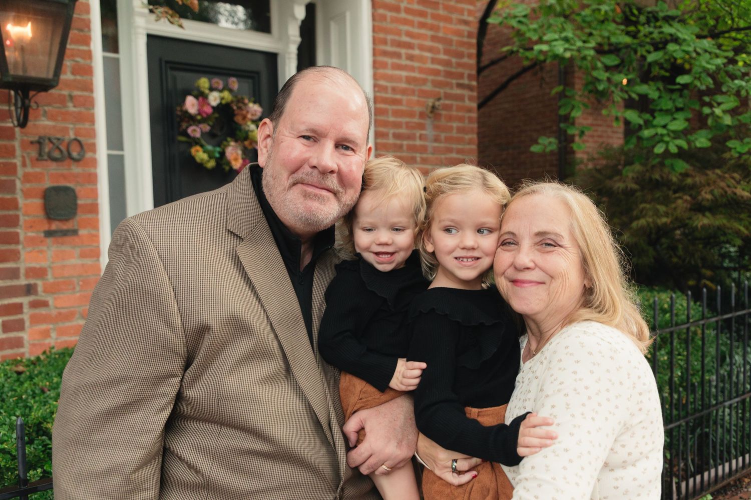 A family of four poses outdoors, with parents holding two young toddlers in front of a brick home entrance.