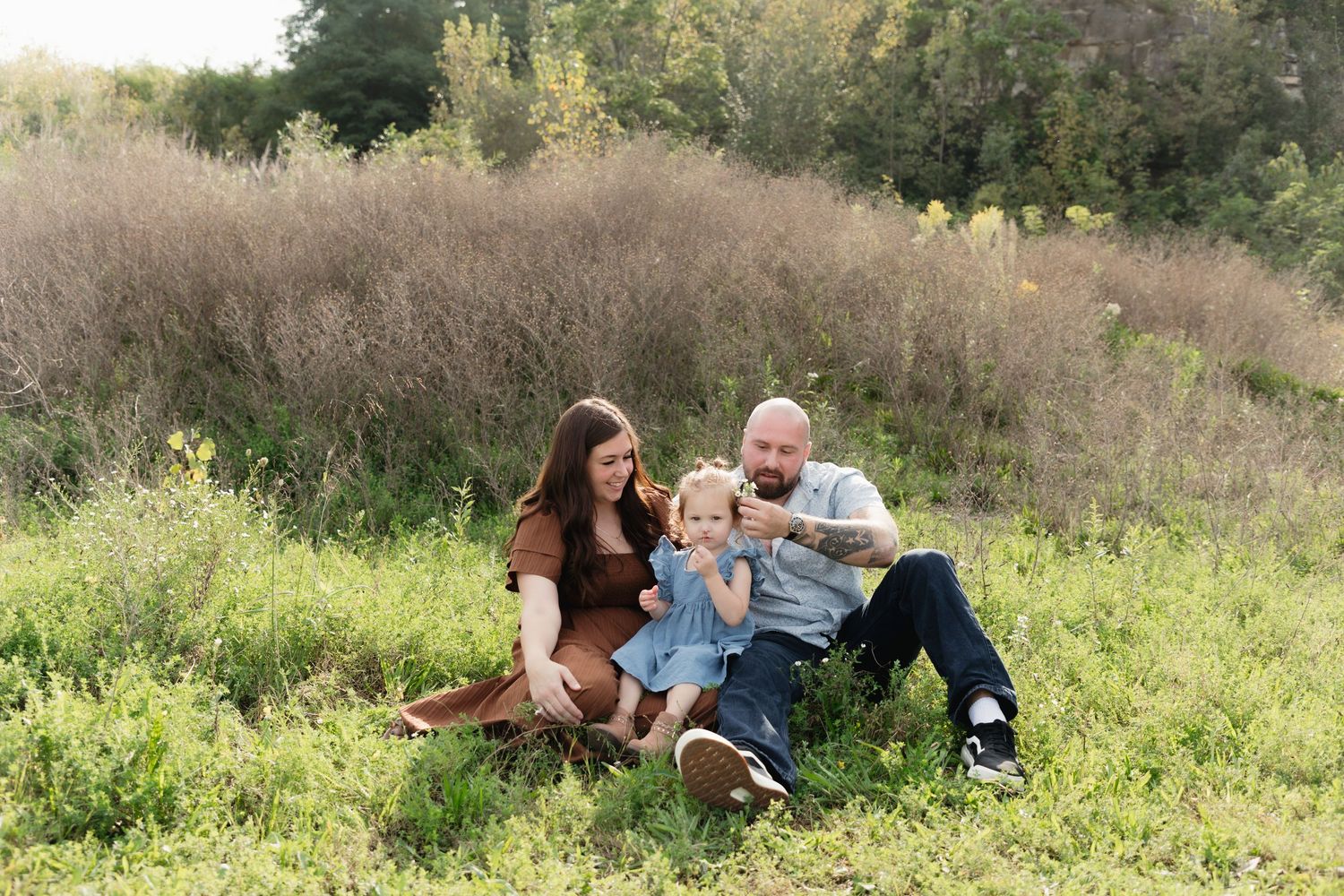 Family of three sitting in a green meadow, parents smiling at toddler on a warm autumn day.
