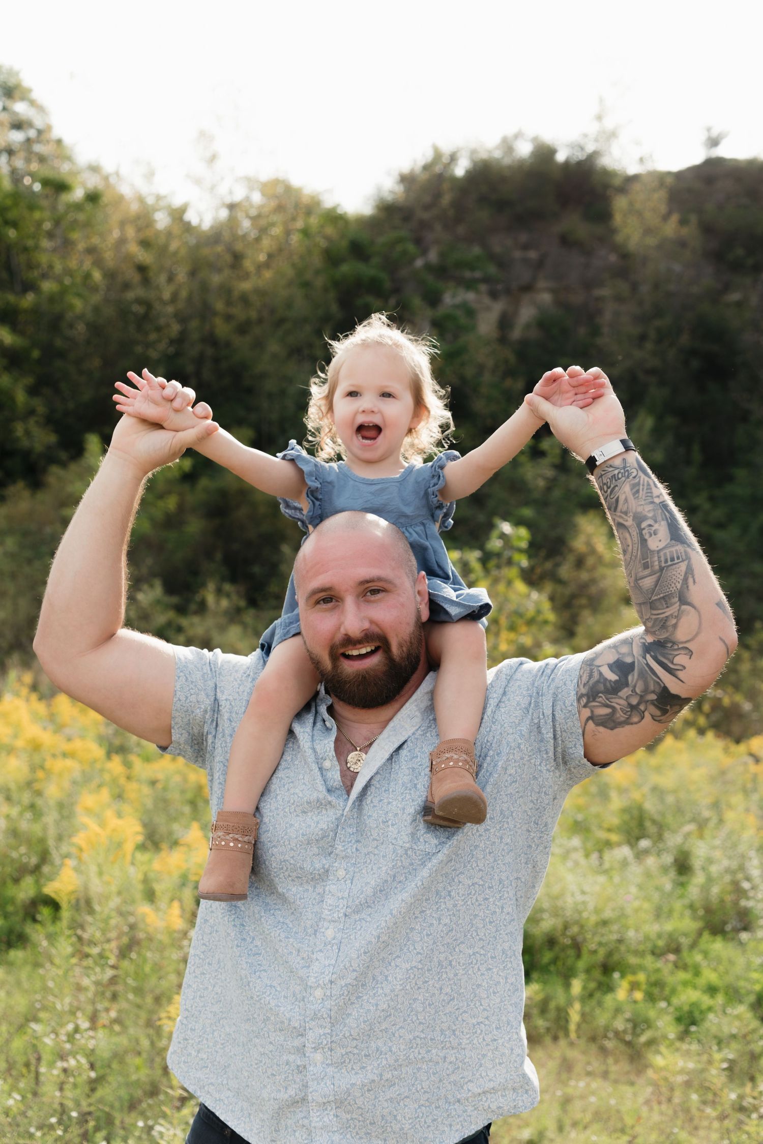 Bald tattooed father holds laughing toddler girl on his shoulders in a sunny outdoor field with green trees.