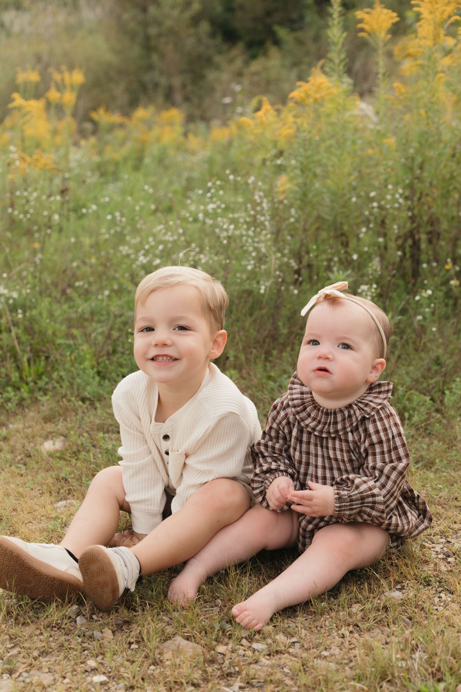Two young siblings sit together in a field of wildflowers, wearing neutral tones, smiling outdoors.