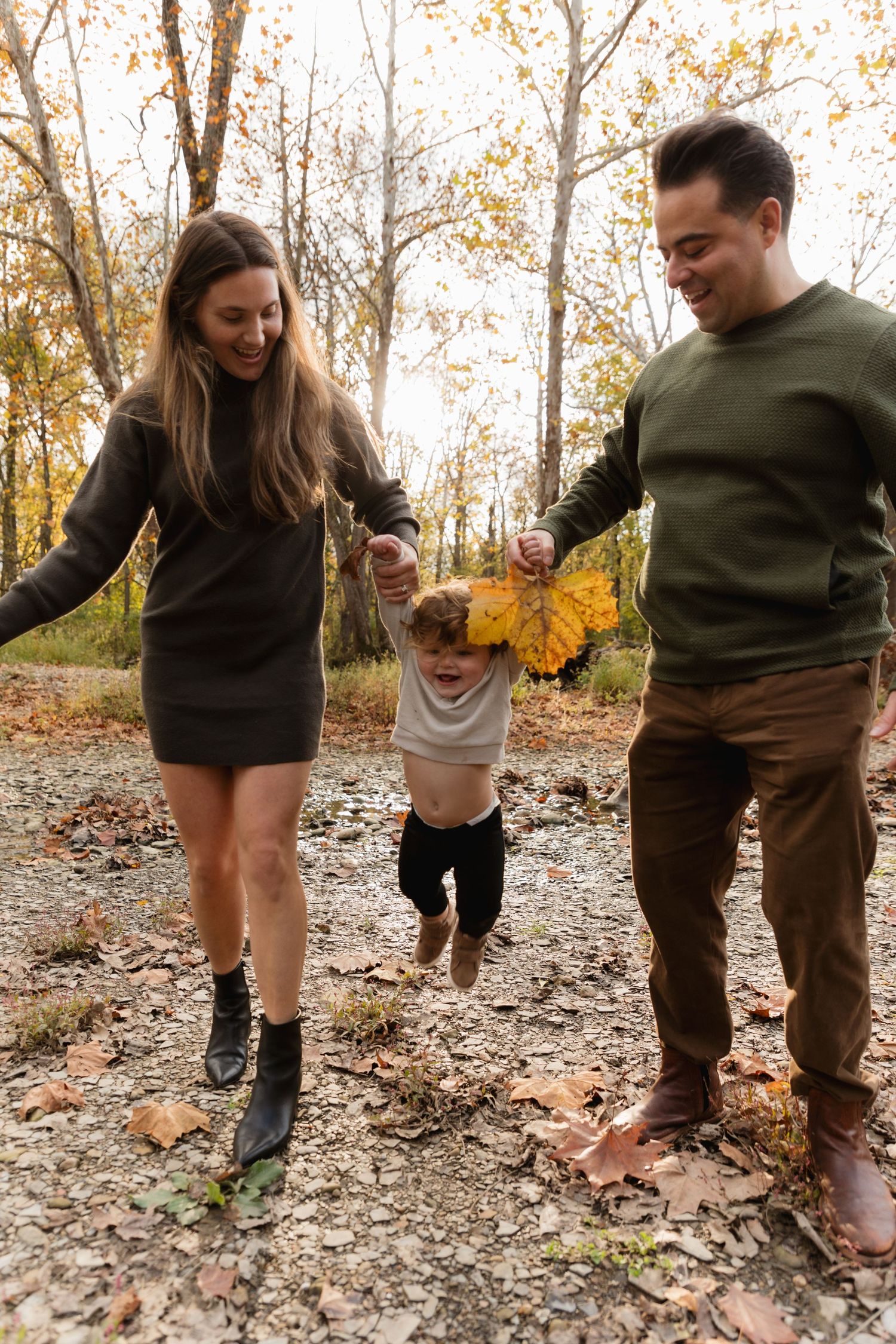 A happy family swings their toddler between them while walking through an autumn forest with golden leaves.