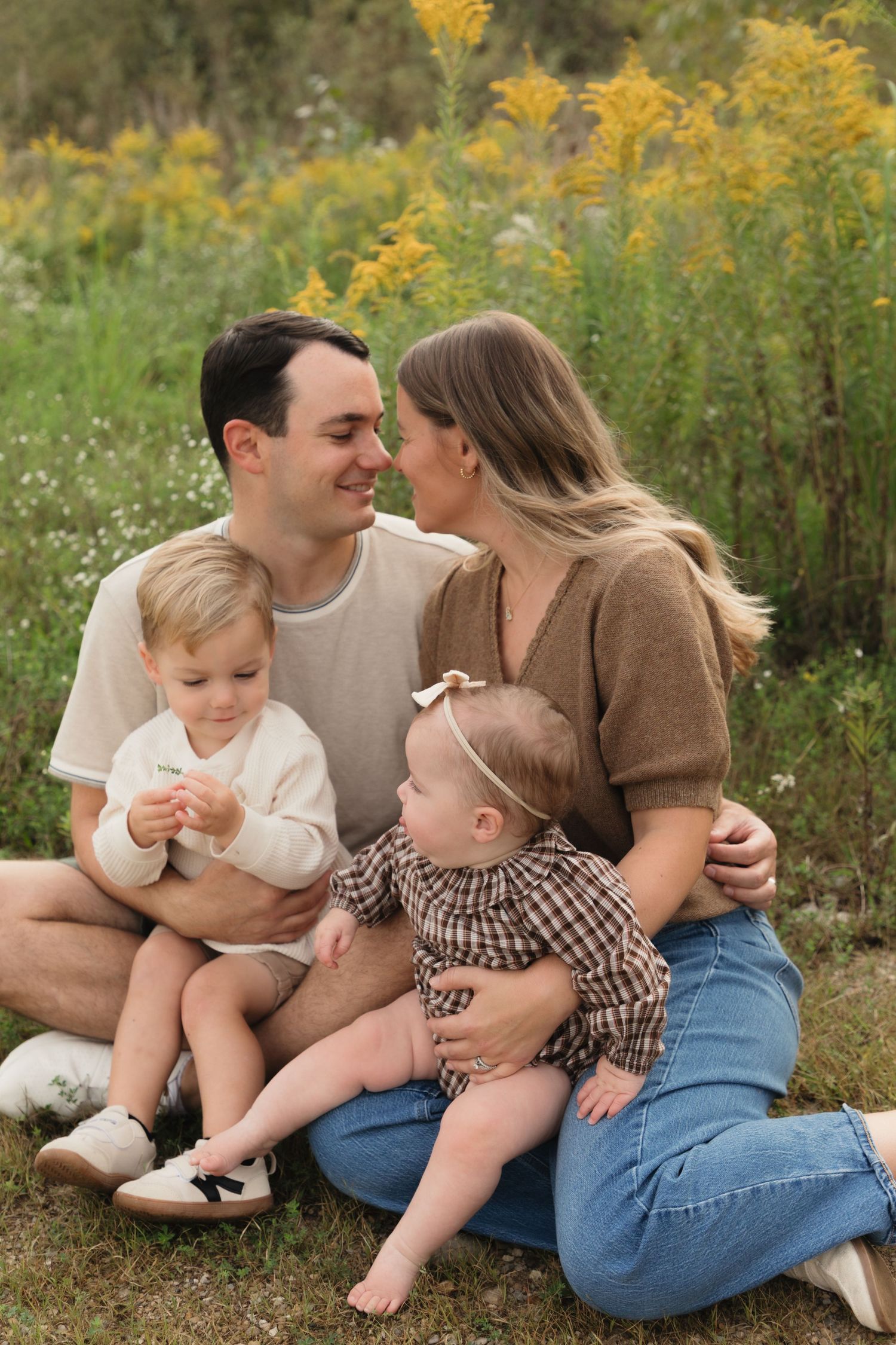 A young family of four sits outdoors in a field of yellow wildflowers, with parents gazing at each other and two toddlers.
