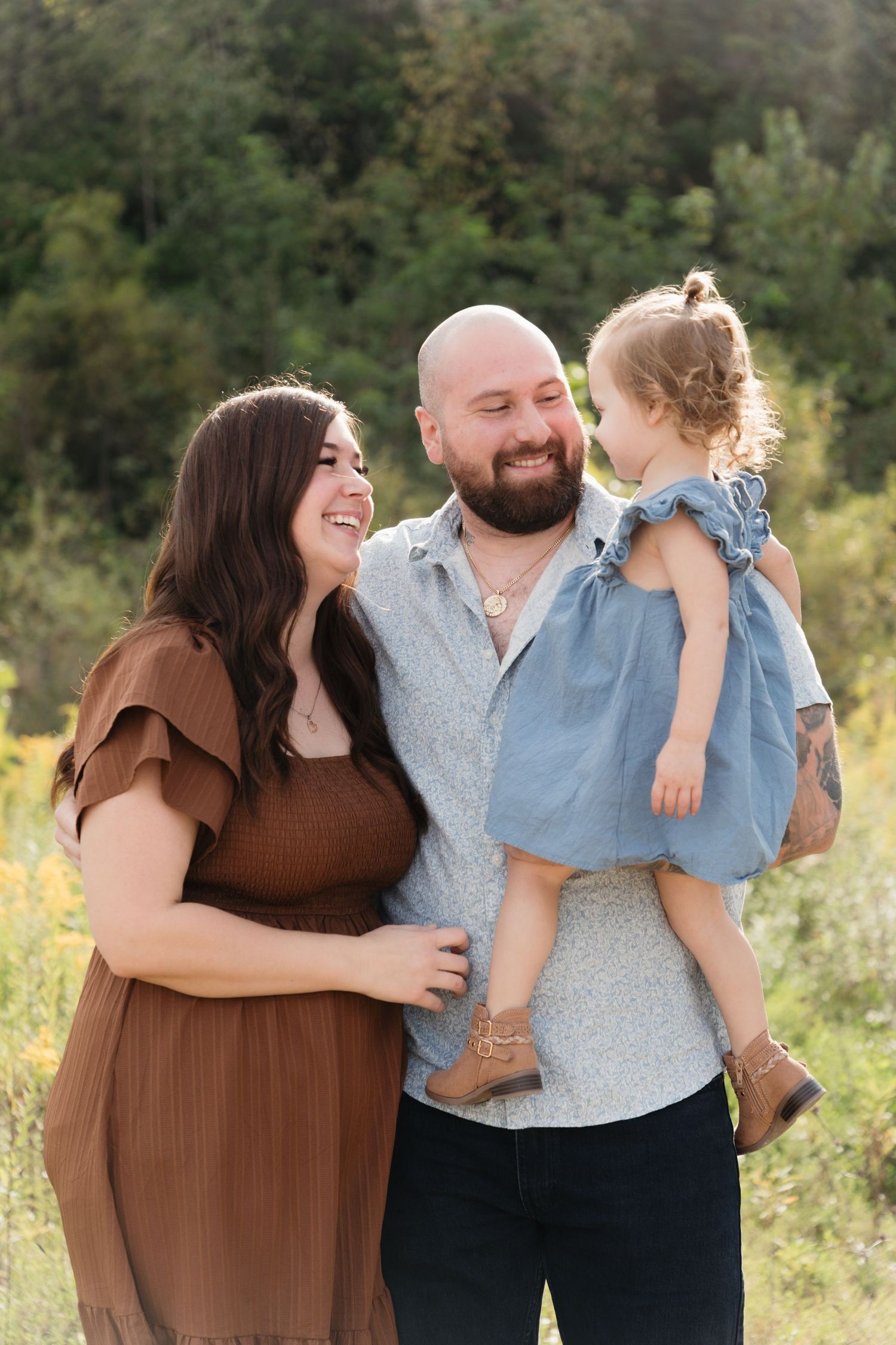Happy family of three posing outdoors, mother in brown dress, father holding toddler girl in blue dress.