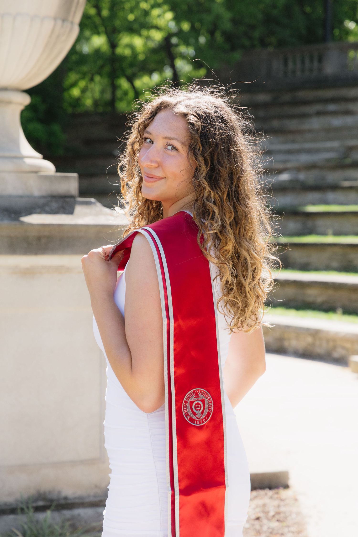 A smiling young woman in a white dress wearing a red graduation sash poses outdoors on a sunny day.