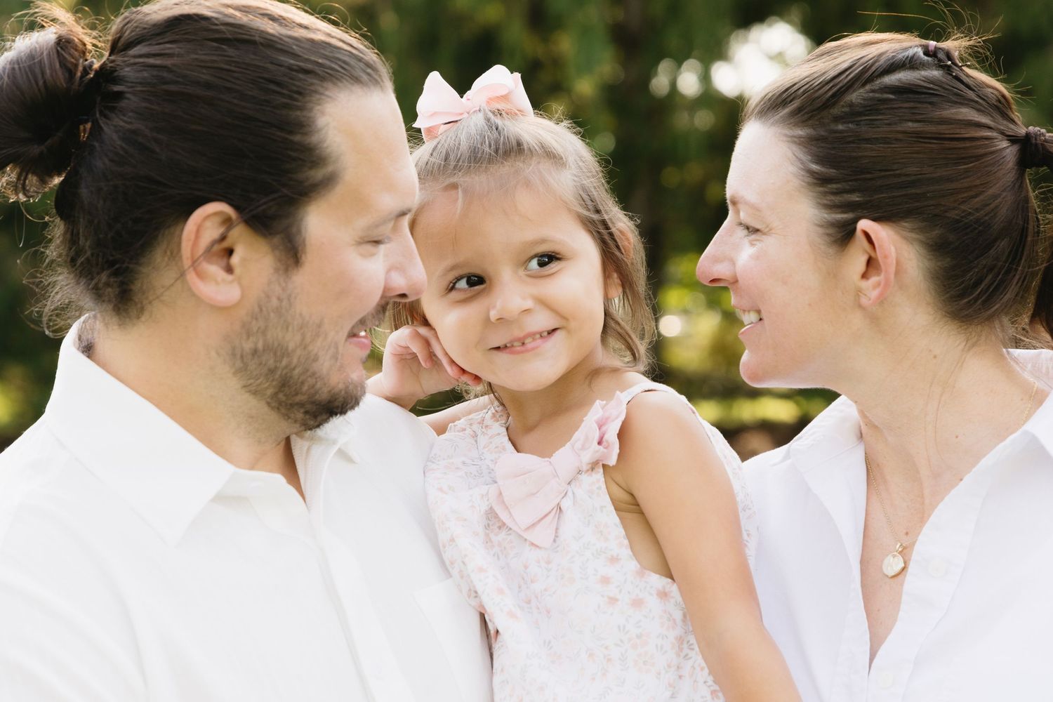 A happy family with parents in white smiling at their young daughter with a pink bow outdoors.