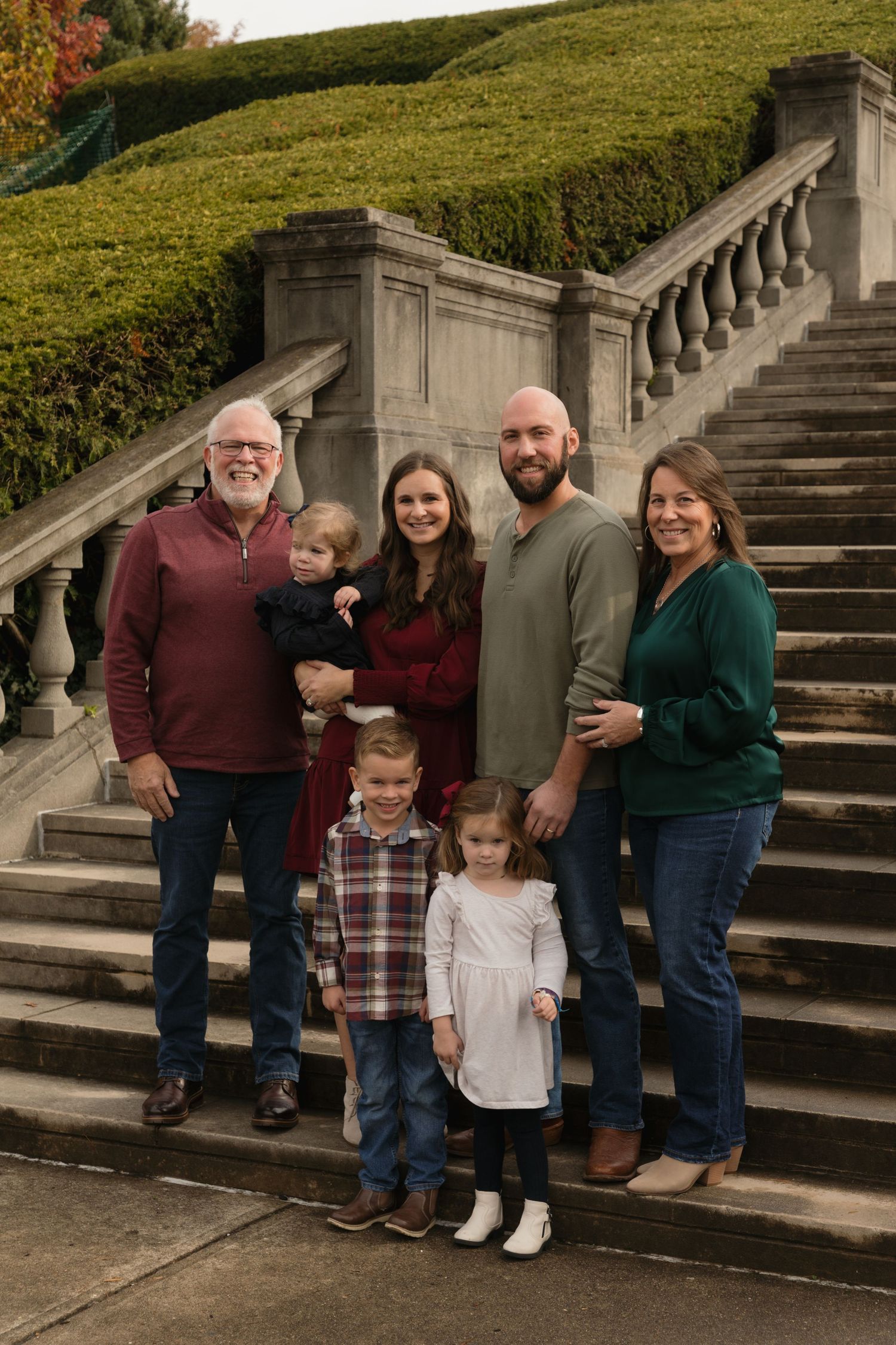 Multi-generational family of seven posing on stone staircase outdoors, dressed in coordinated fall colors for a portrait.