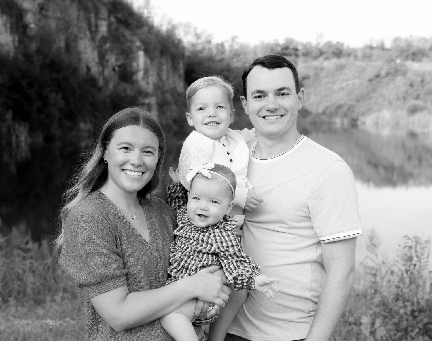 Black and white family portrait of smiling parents holding two young children outdoors near a river.