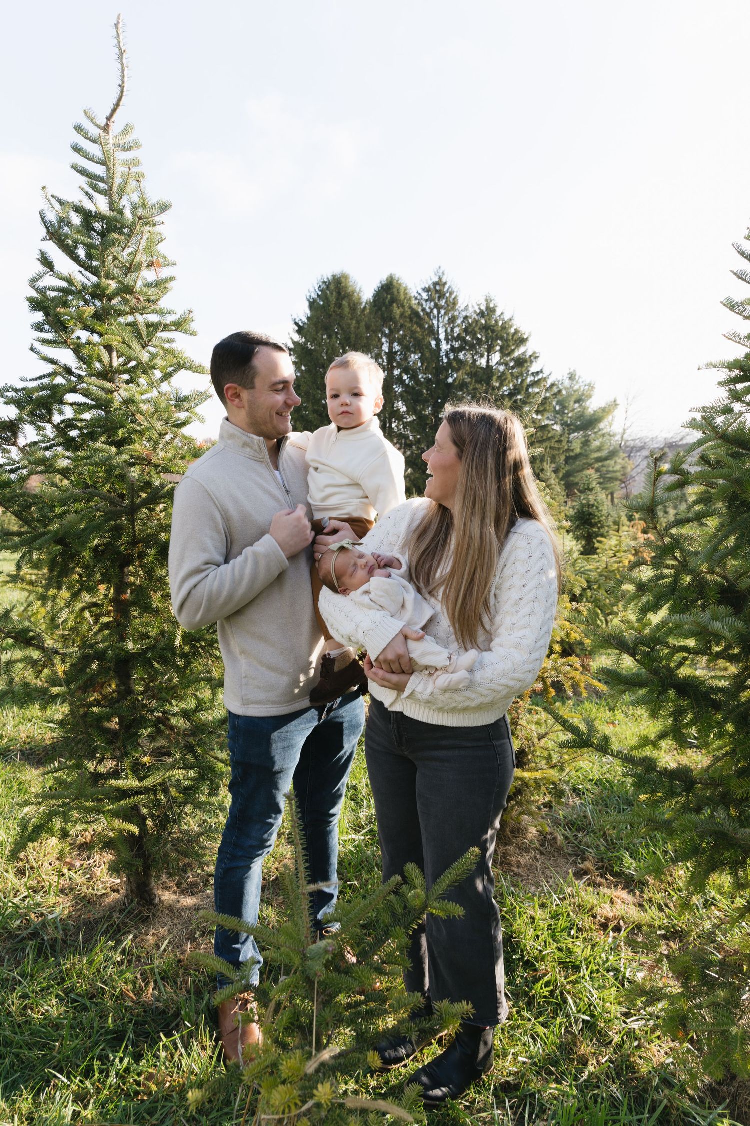 A happy family with a baby poses among green Christmas trees at a tree farm on a sunny day.