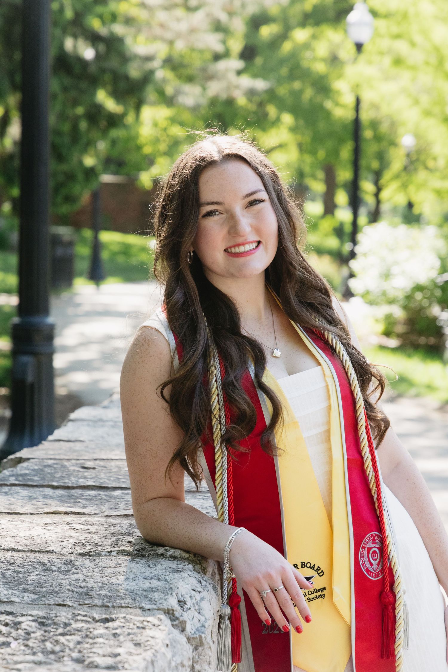 Smiling college graduate in red and gold stole poses outdoors on a sunny campus walkway.