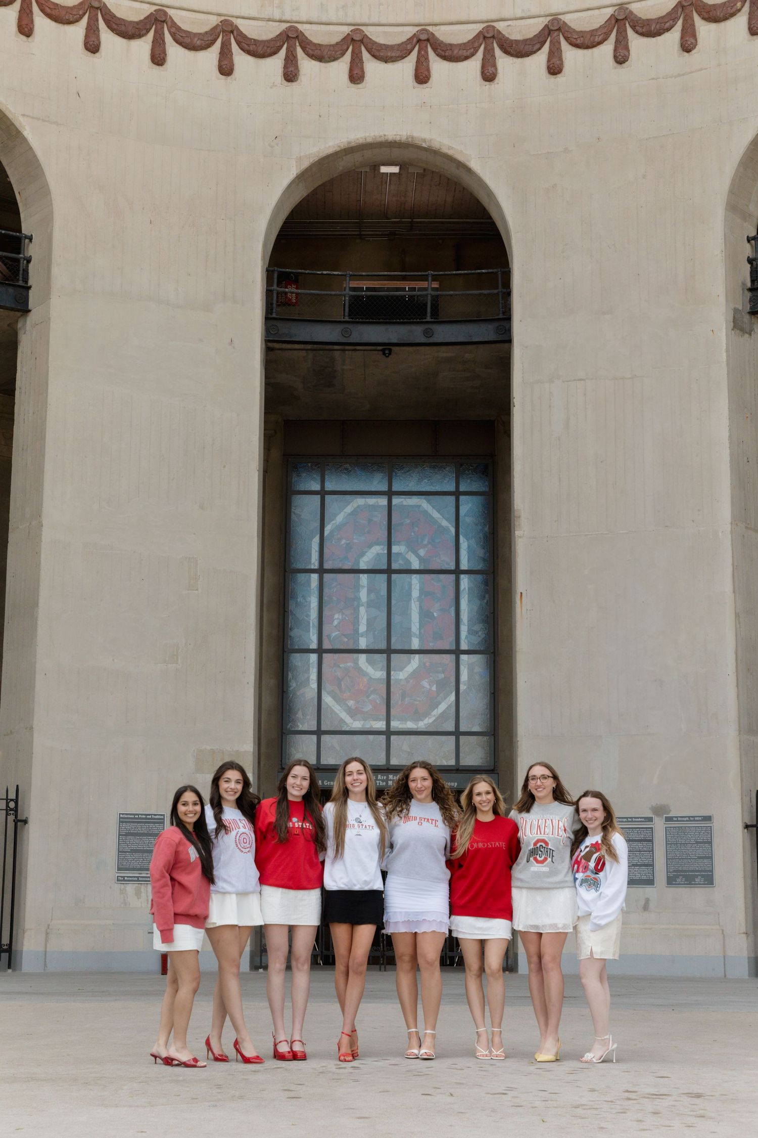Eight young women in red and white outfits pose together outside Ohio State University's iconic stadium archway.