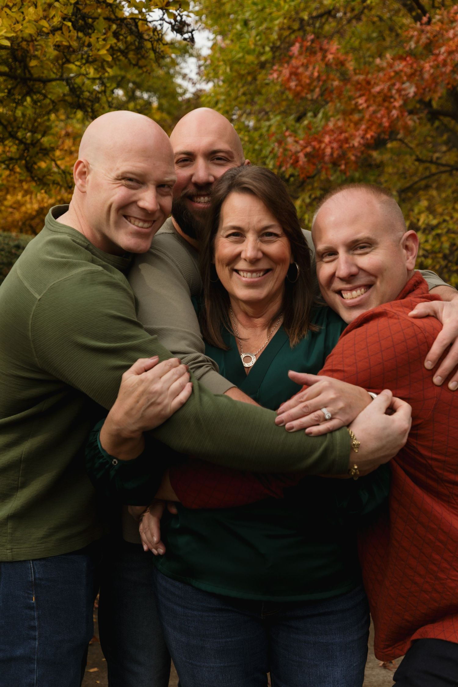 Four smiling adults in fall foliage, embracing outdoors in autumn colors, wearing green and red tops.