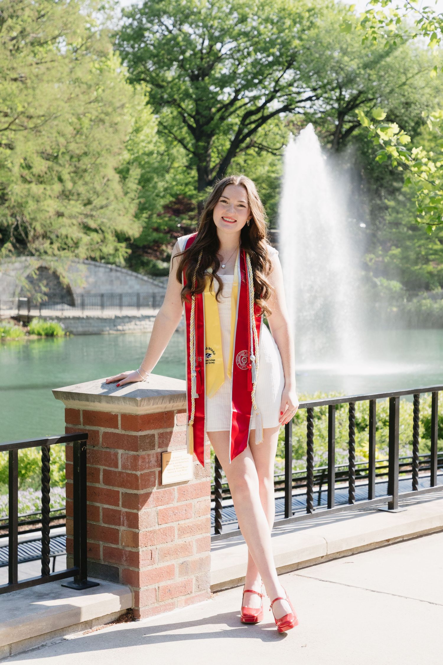 Smiling graduate in white dress with red and gold stole poses by fountain on sunny campus day.