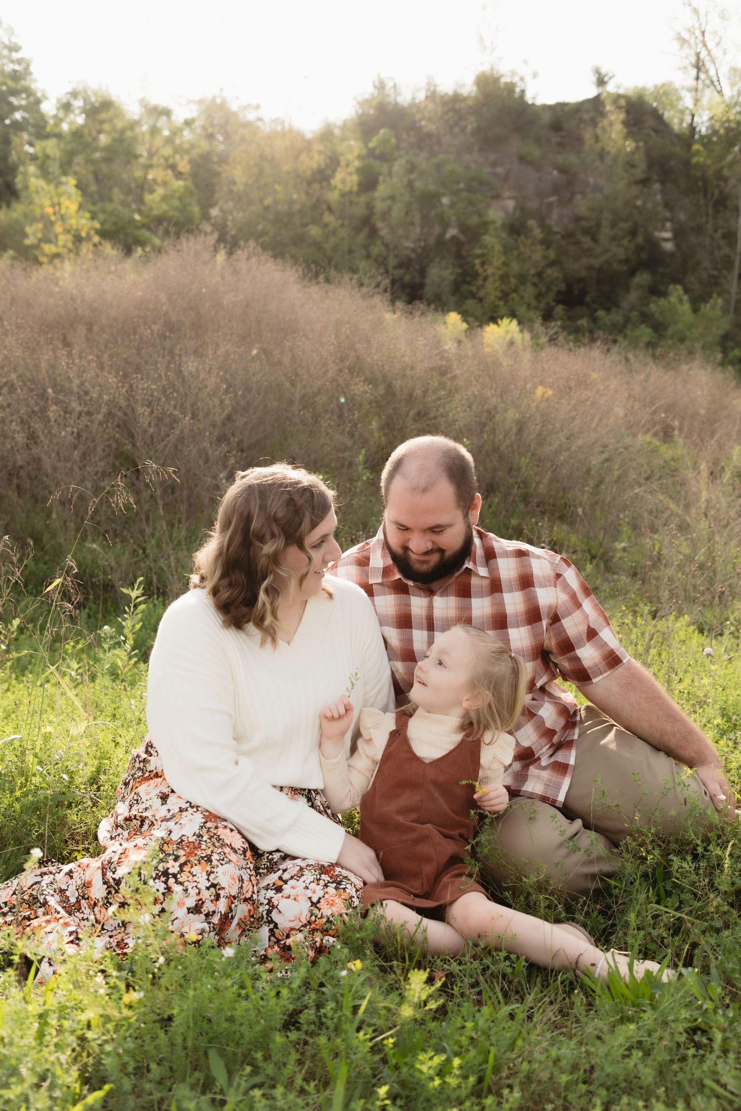A family of three sits together in a sunlit meadow, parents smiling at their young daughter in a rust-colored dress.