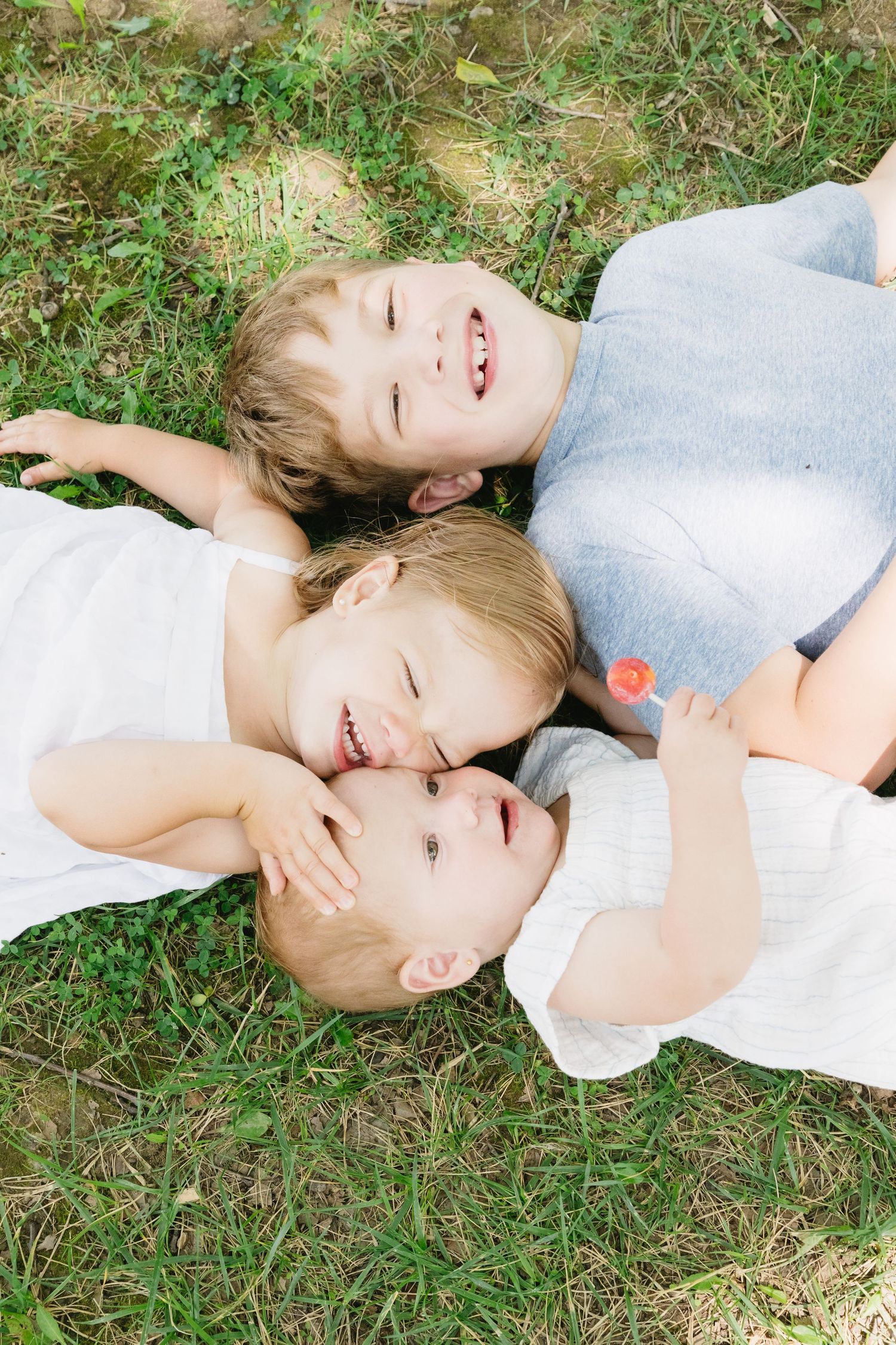 Three happy children lying on green grass together, smiling and cuddling in a heartwarming outdoor sibling portrait.