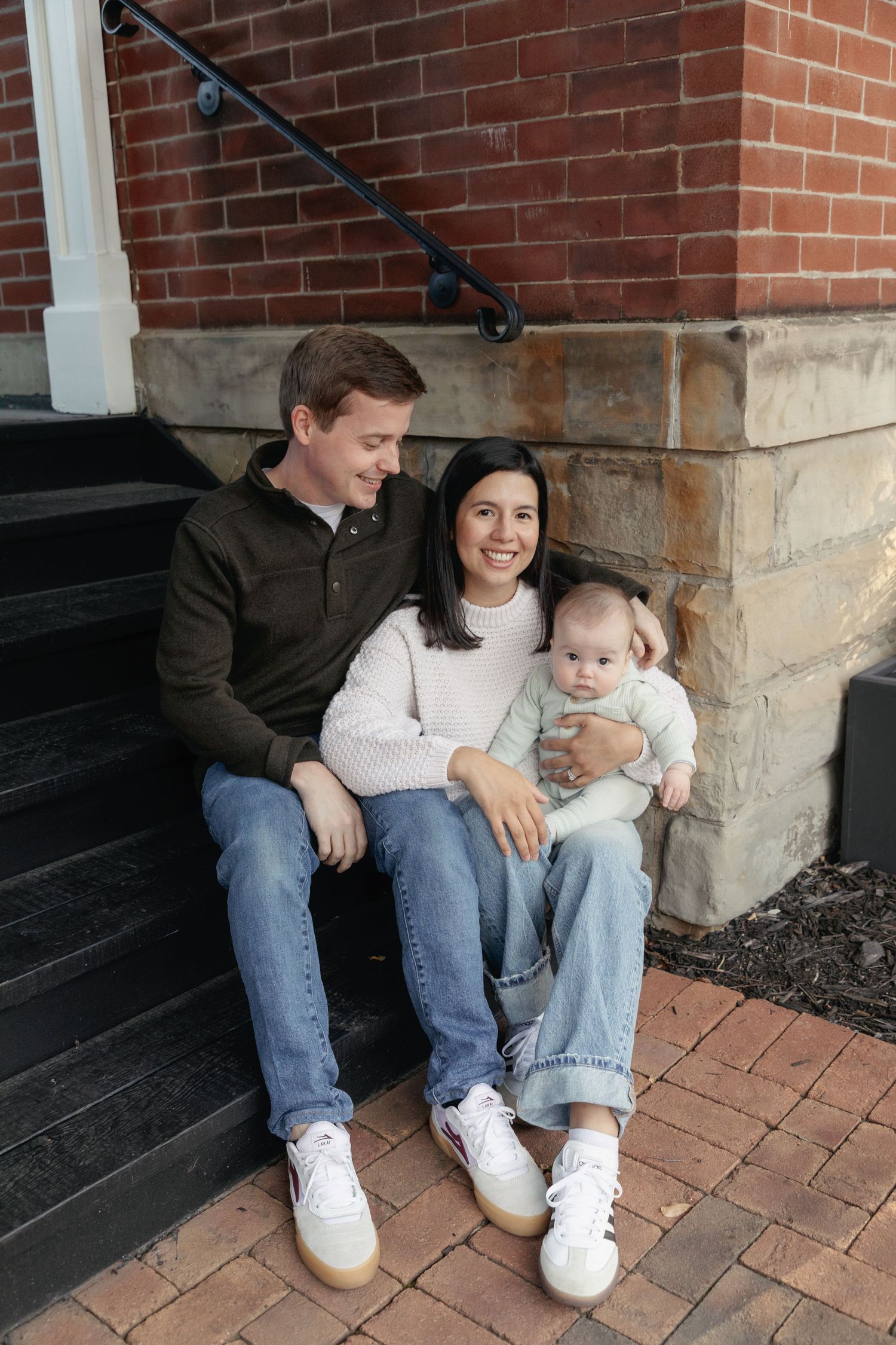 Young family with baby sitting on brick front porch steps of a home, smiling and enjoying time together.