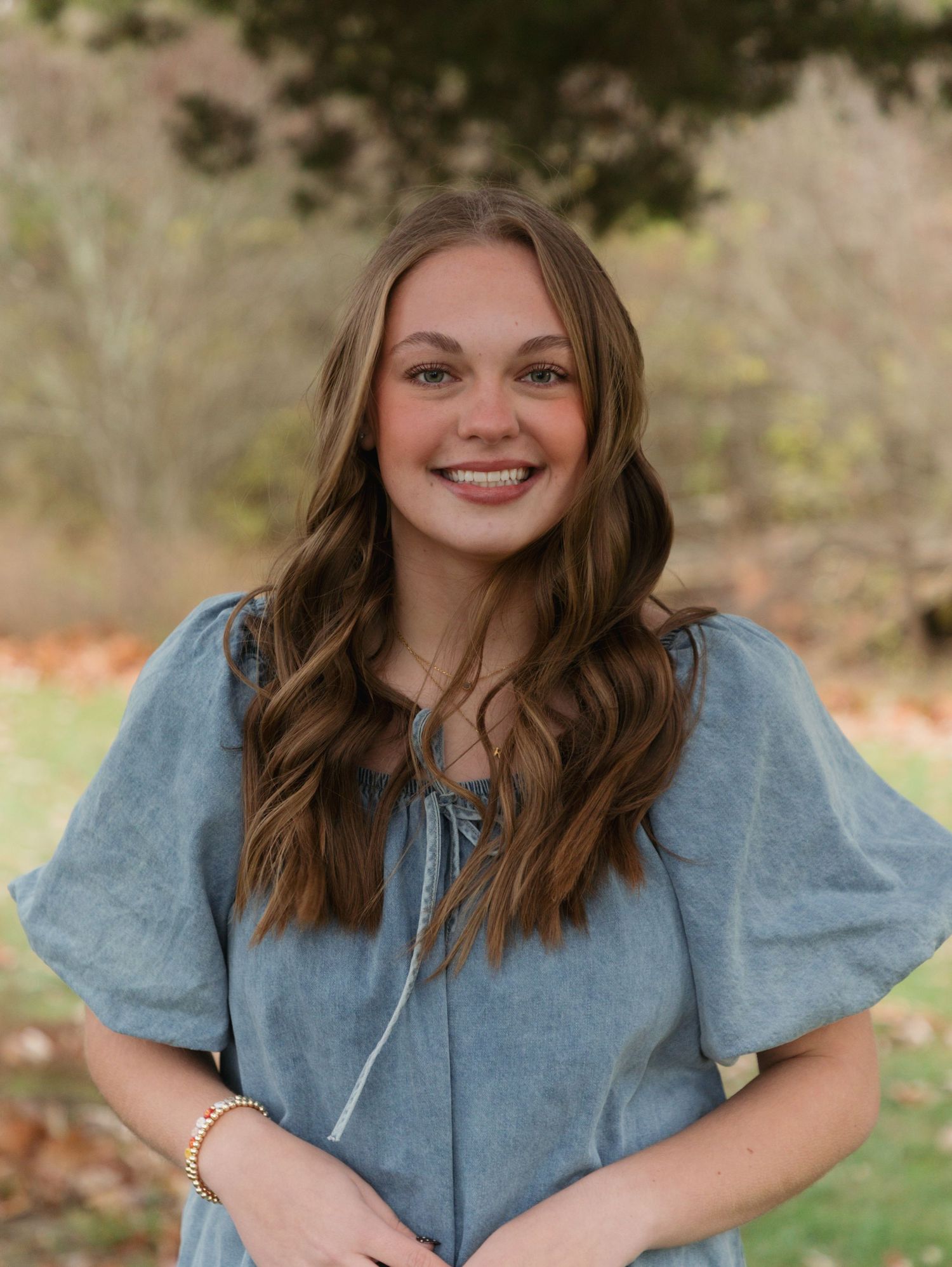Young woman with long brown wavy hair smiling outdoors in a blue dress, with a natural green background.