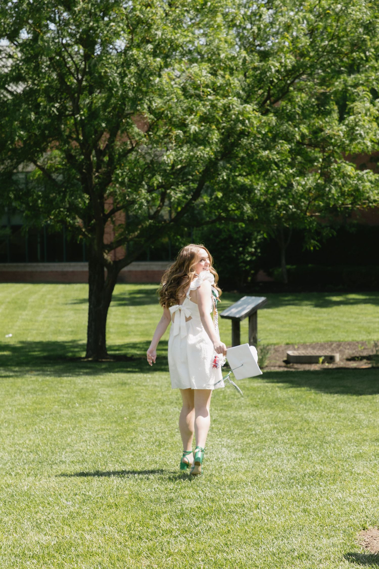 Young woman in white dress holding graduation cap walks across green campus lawn on a sunny day.