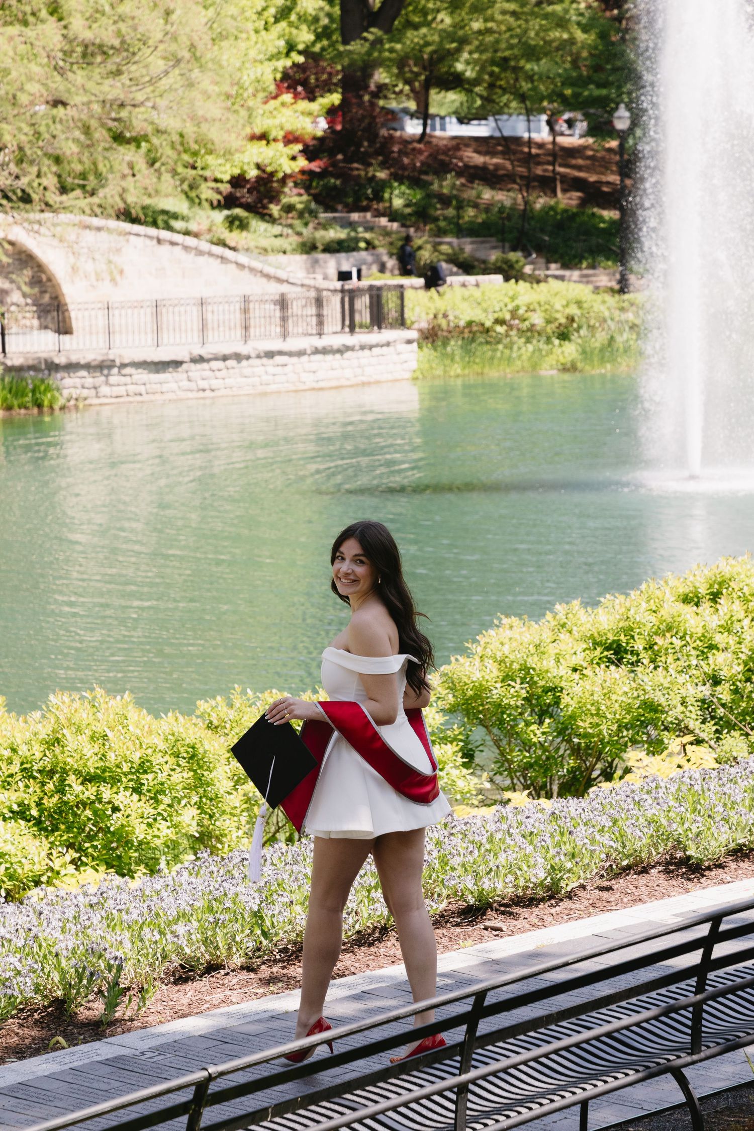 Young woman in white off-shoulder dress holding graduation cap by a fountain and green garden.