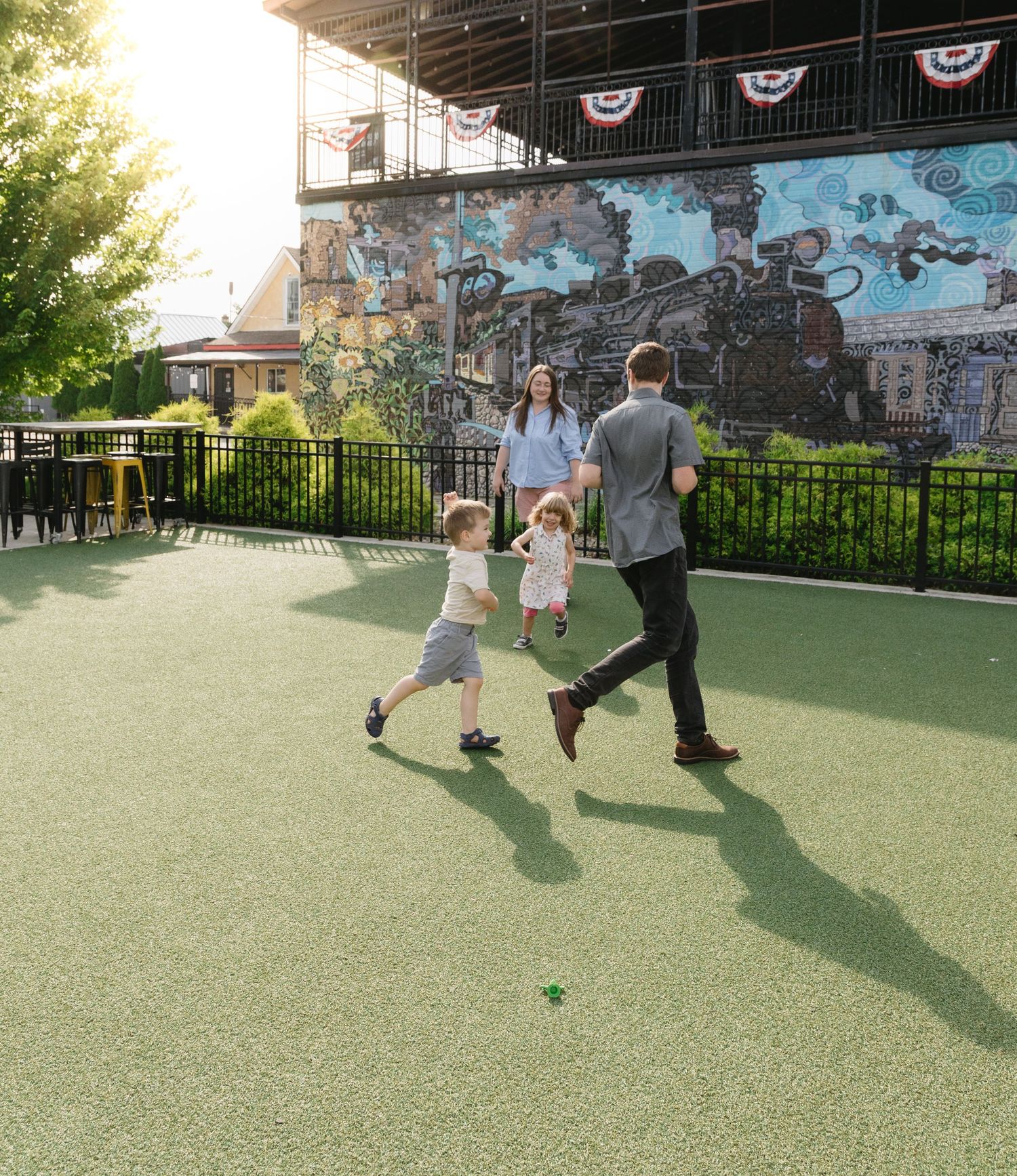 Family playing outdoors on green turf near a colorful train mural at a vibrant outdoor venue.
