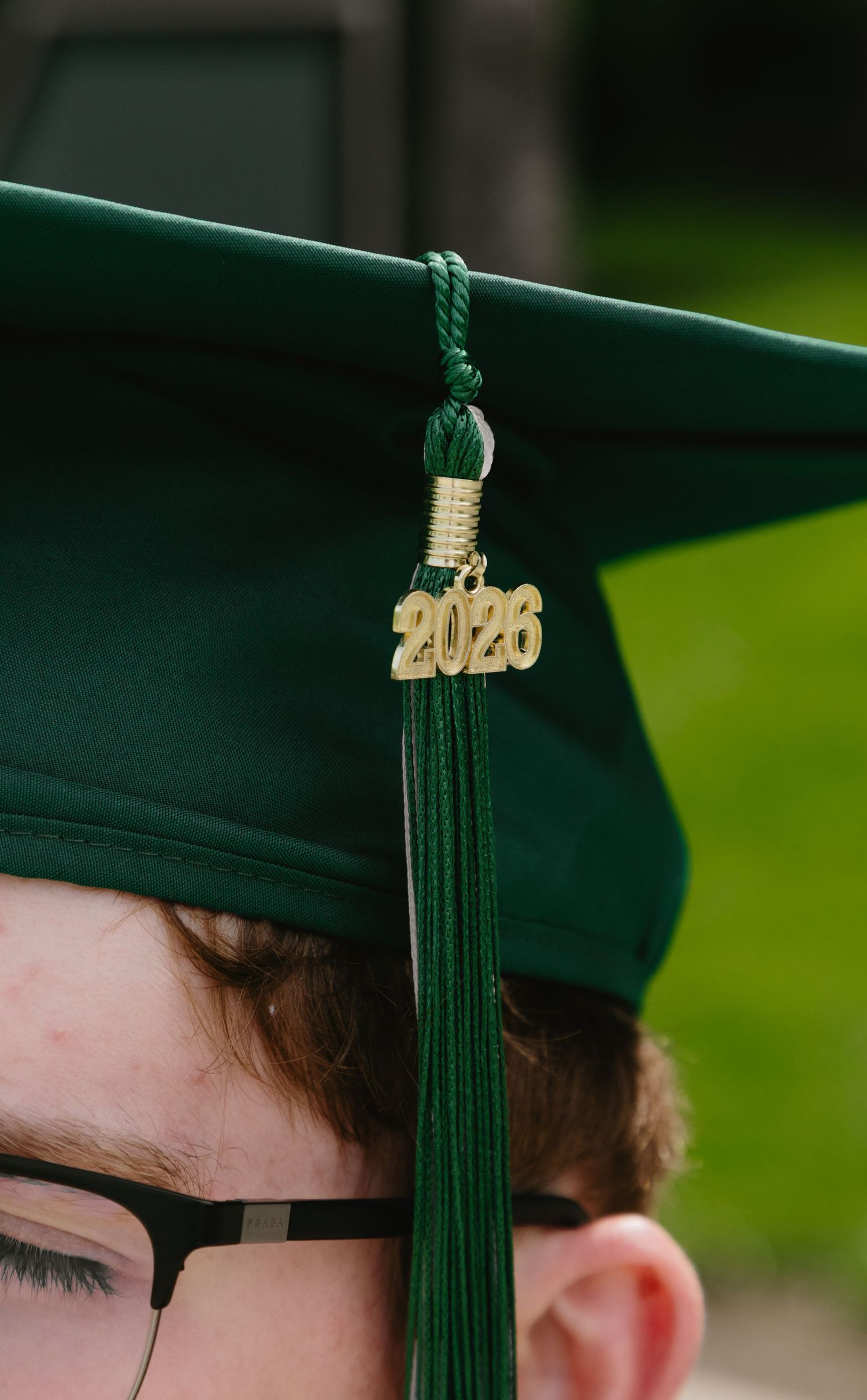 Close-up of a green graduation cap with a gold 2026 tassel charm worn by a student with glasses.