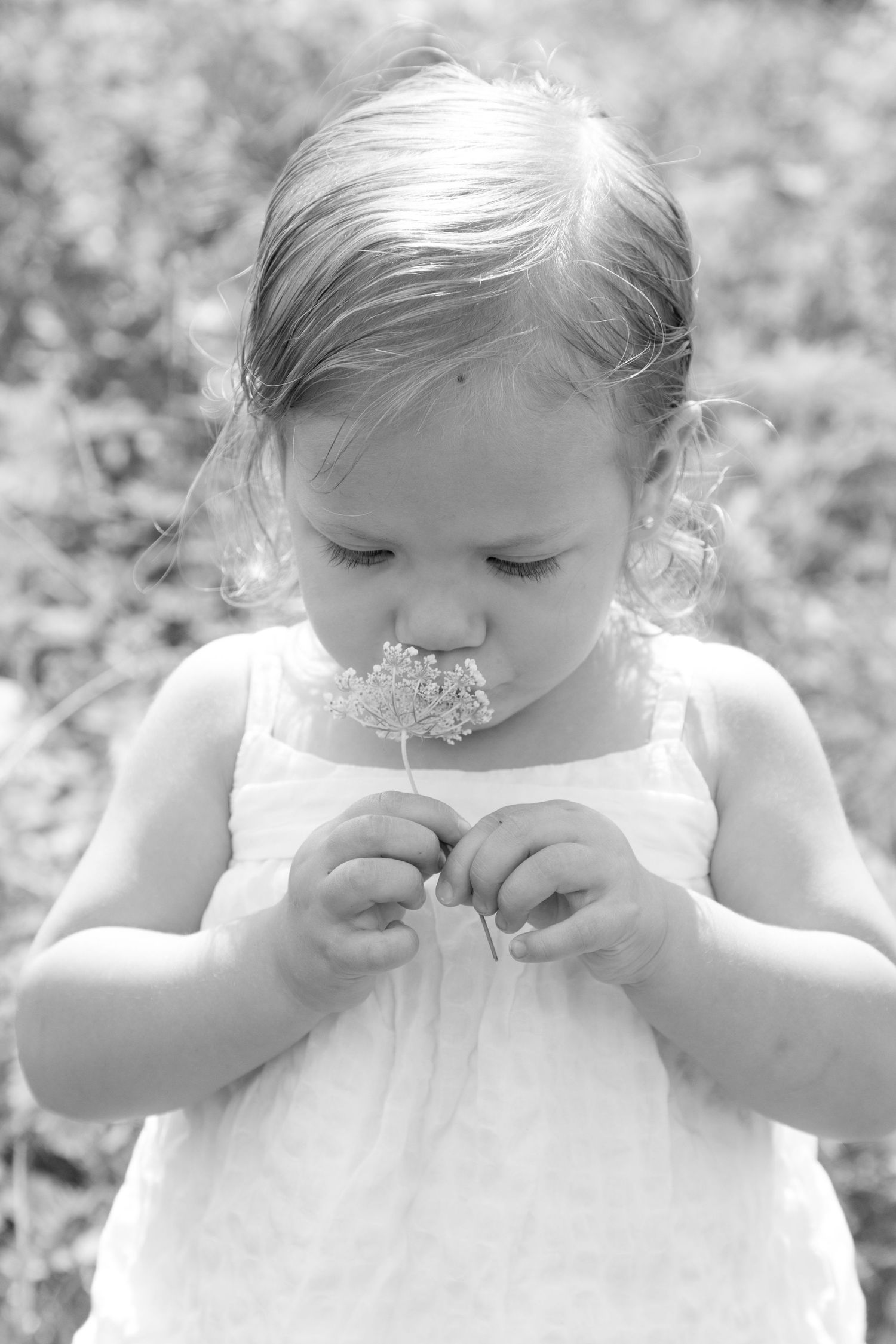 Black and white photo of a young girl in a white dress smelling a small wildflower outdoors.