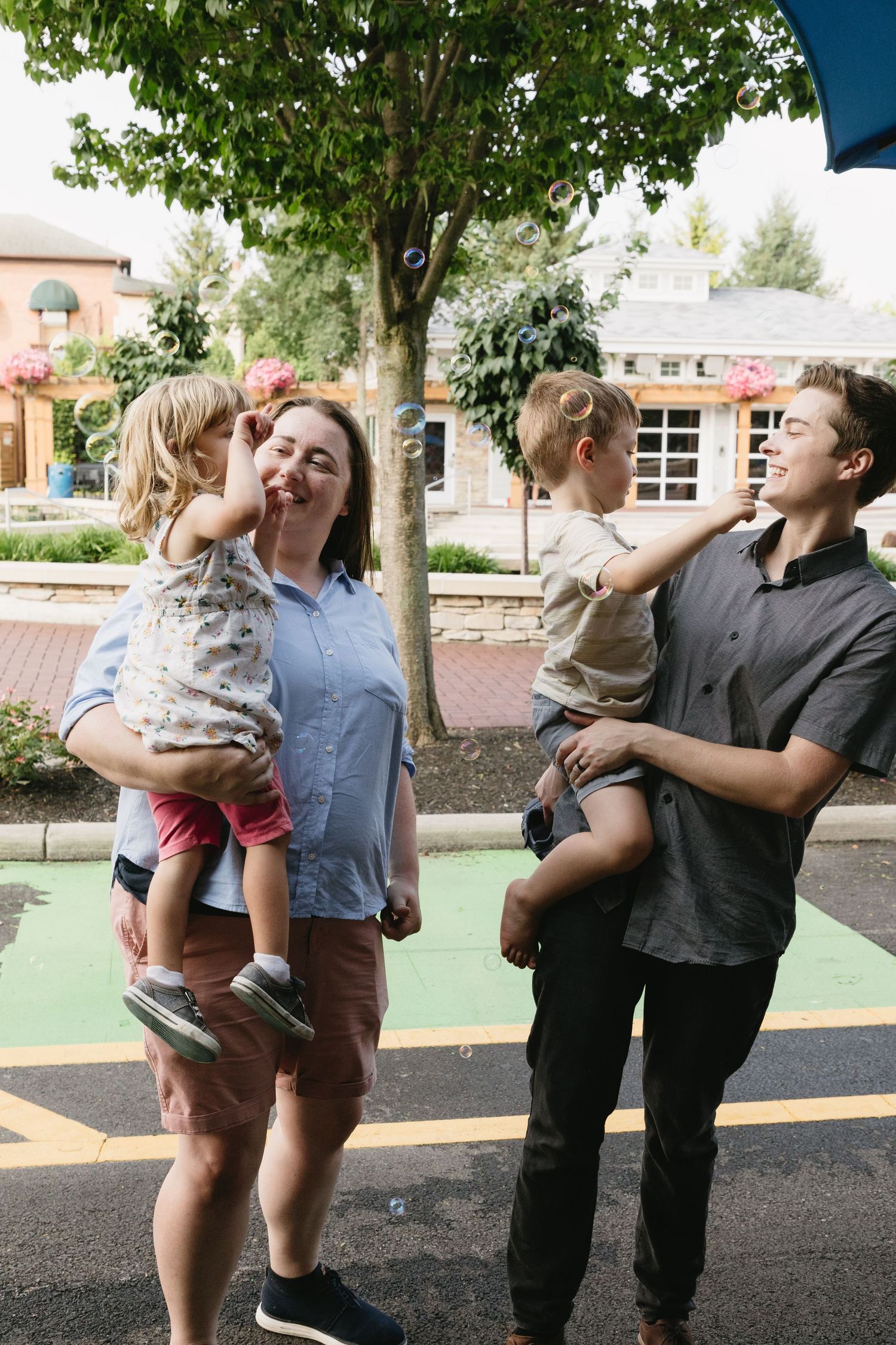 A family of four with two young children being held by parents outdoors on a street, smiling and playing together.