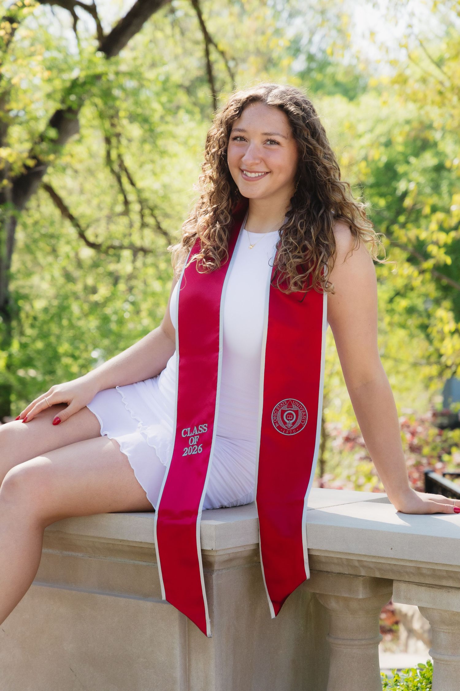 Young woman in white dress wearing red graduation sash smiles outdoors among green trees on a sunny day.