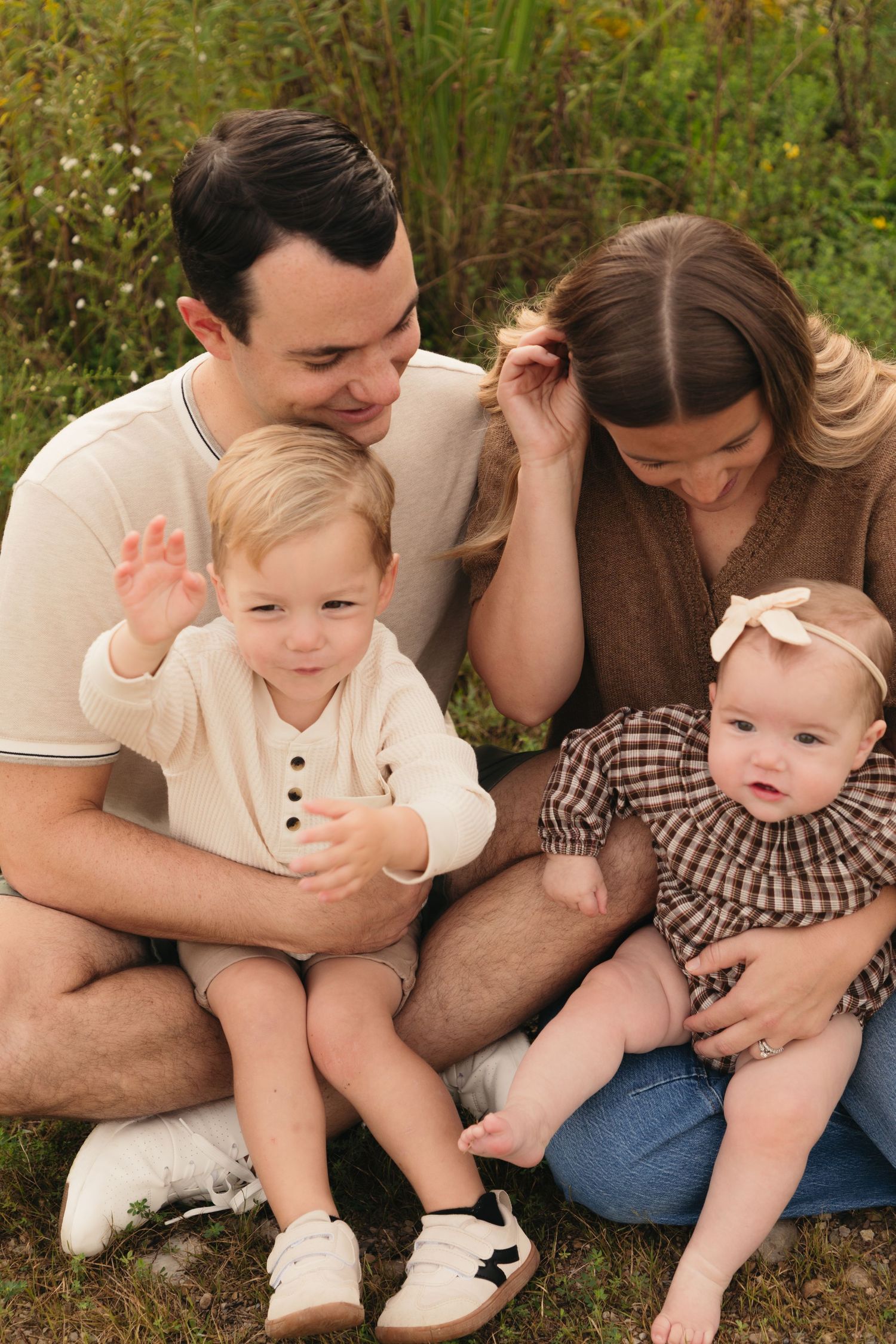A young family of four sits outdoors on grass, with two parents smiling at their toddler and baby girl in a plaid dress.