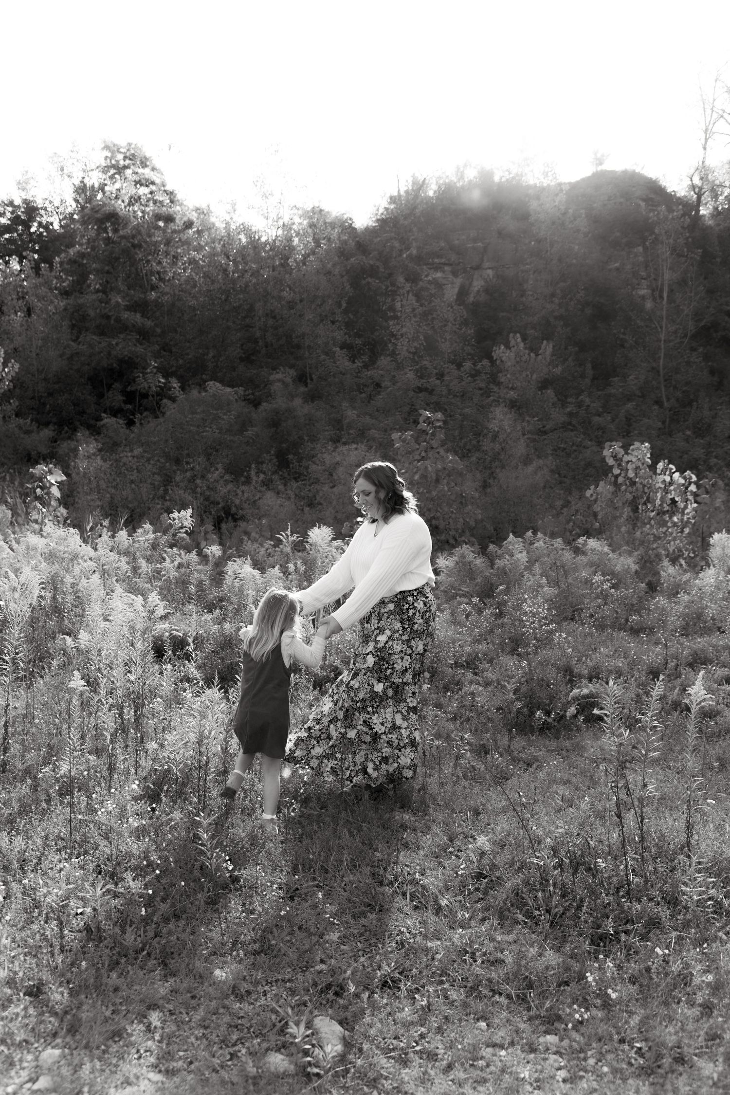 Black and white photo of a mother and young child holding hands in a lush meadow with trees in the background.
