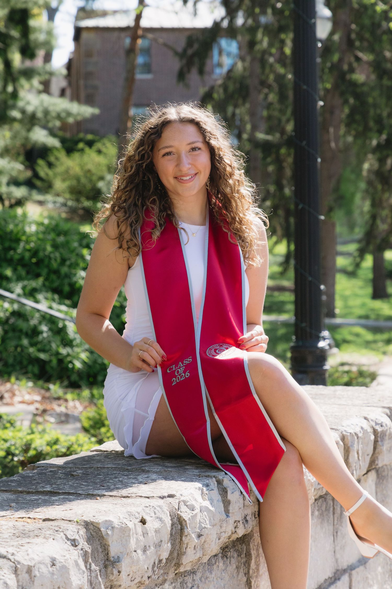 Young woman in white dress and red graduation stole sits on stone wall outdoors, smiling for senior photos.