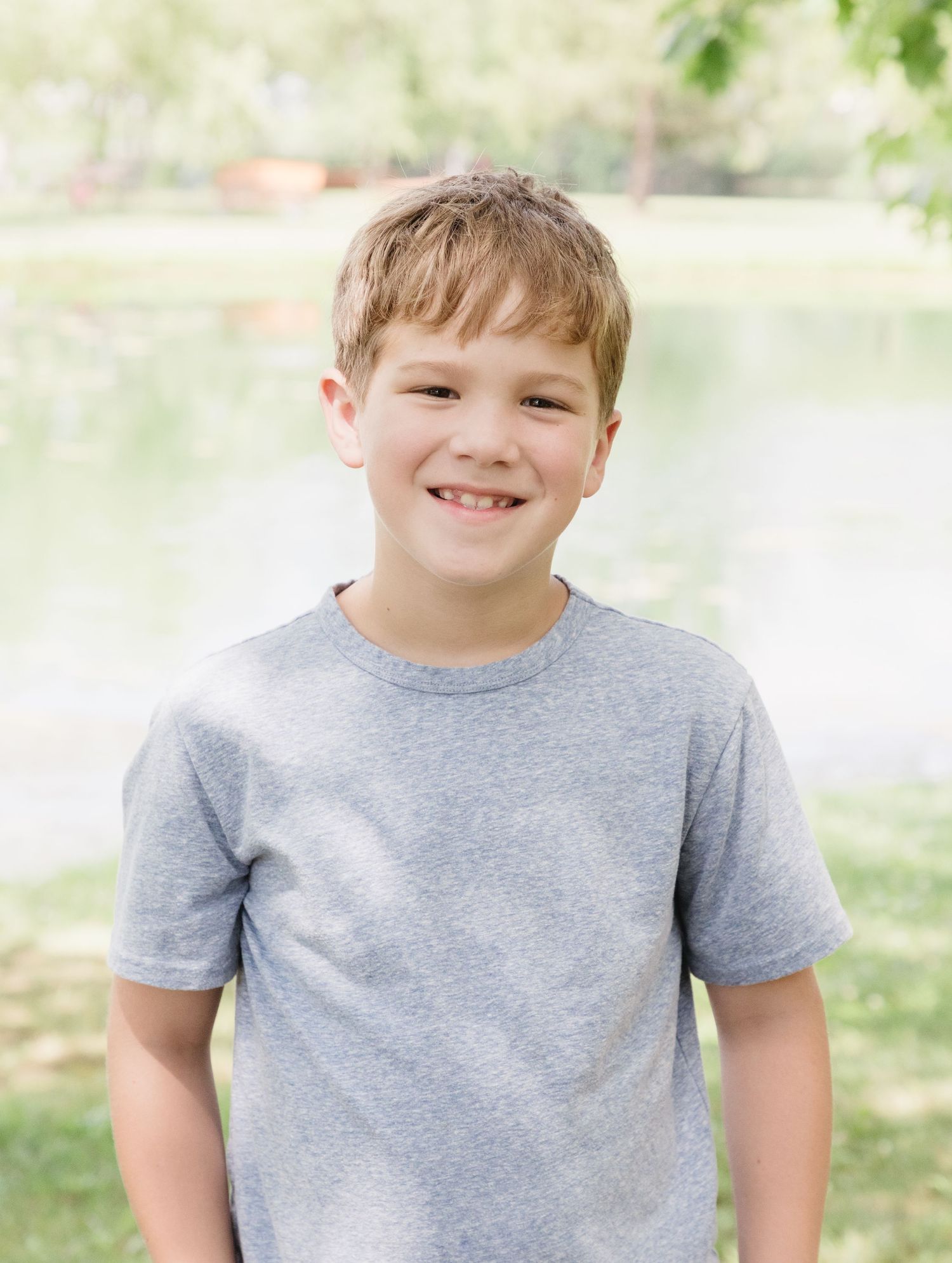 Smiling young boy in a gray t-shirt posing outdoors near a pond with green trees in the background.