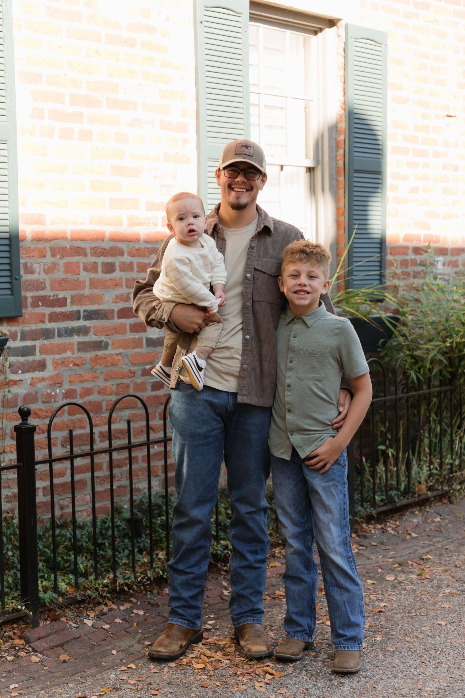 A man in a cap holds a baby while standing with a young boy in front of a brick building.