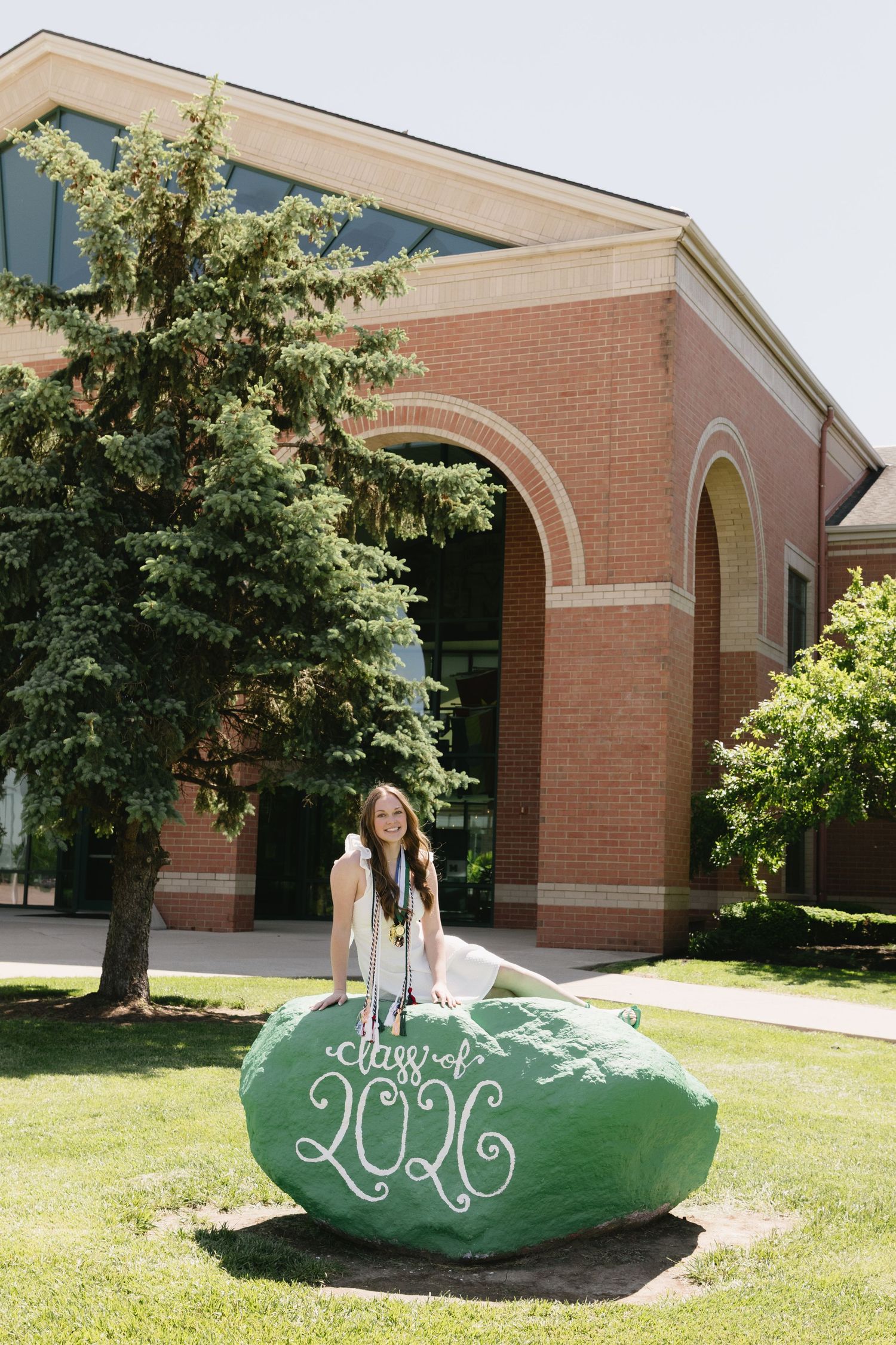 A graduate poses on a green painted rock reading Class of 2026 in front of a brick university building.